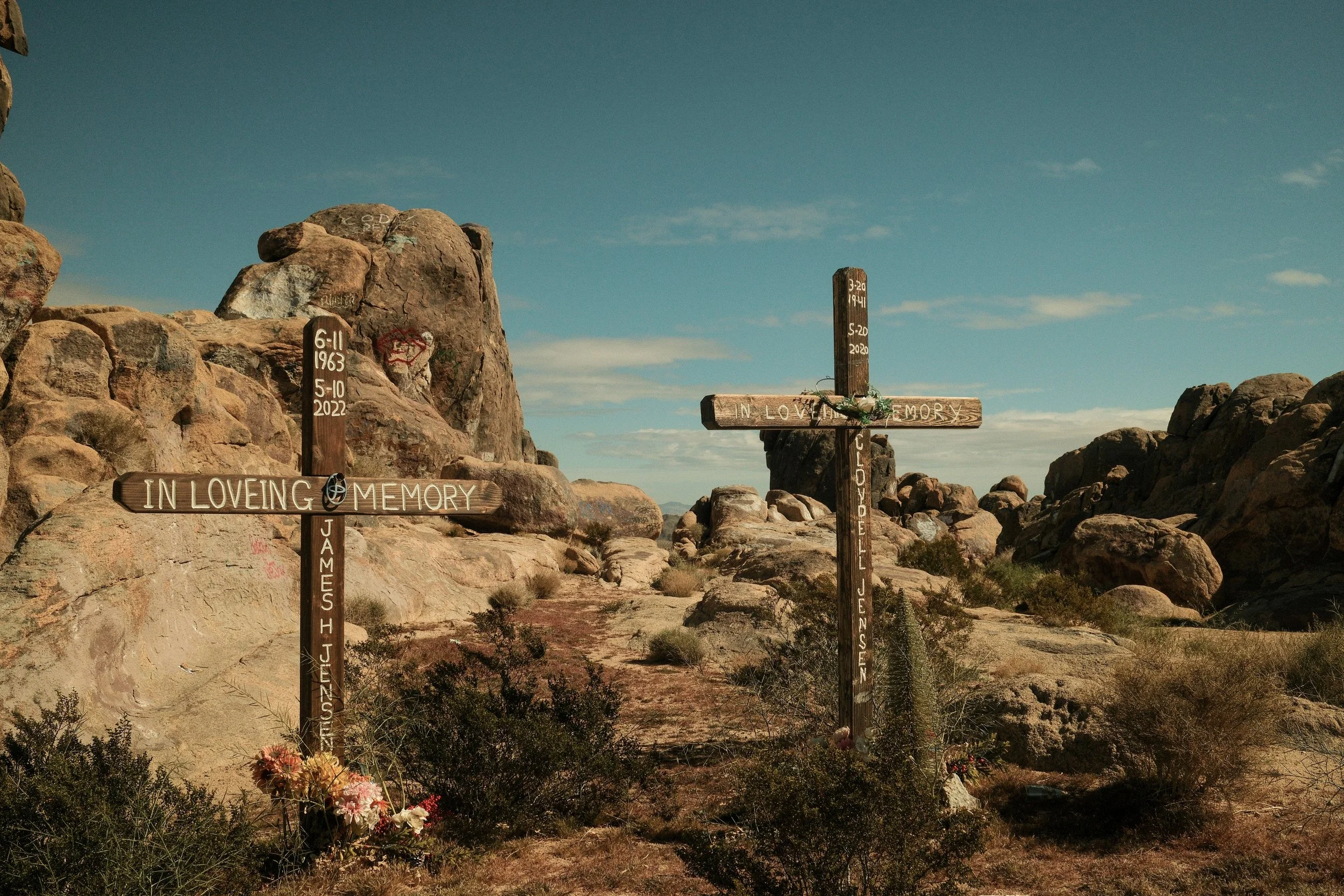 Two wooden crosses with inscriptions, placed in a desert landscape with rocks and sparse bushes under a blue sky, memorializing James H. Jensen and El Jessen, with dates and personal messages.