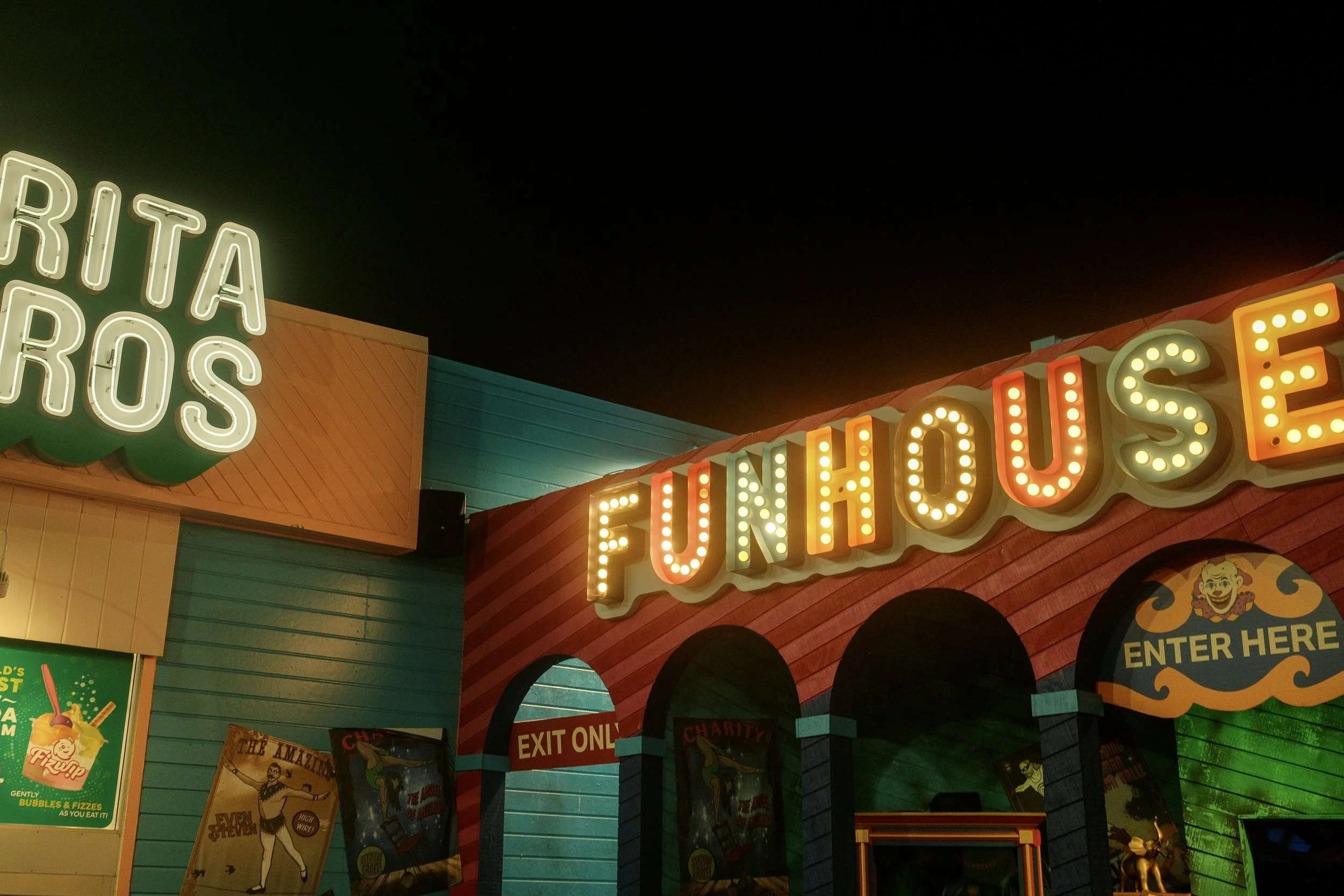 Night view of a carnival or amusement park arcade entrance with brightly lit signs that say 'FUNHOUSE' and 'ARITA ROS', colorful structures, poster advertisements, and an 'ENTER HERE' sign with a clown face.