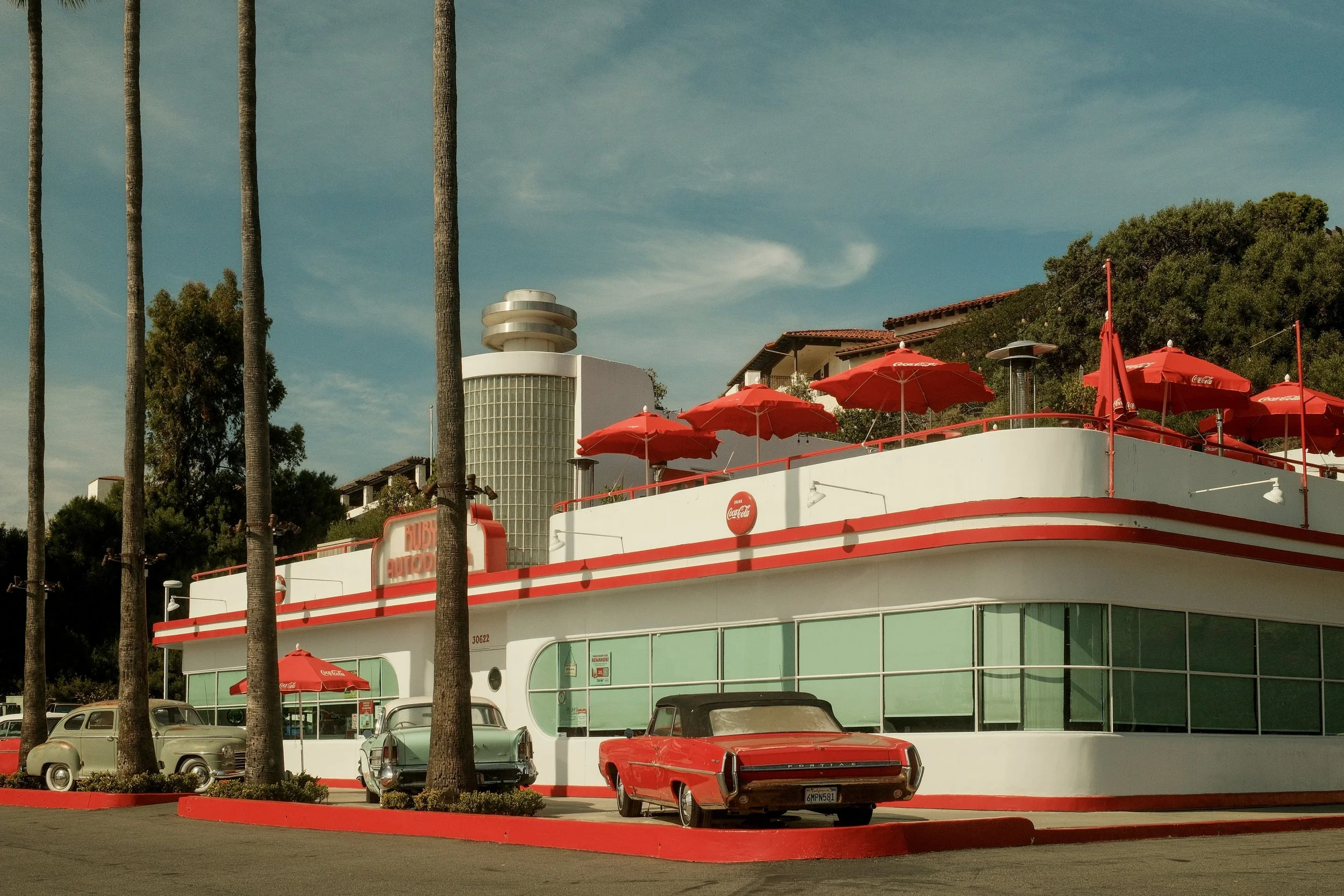 Retro diner with white walls and red accents, outdoor seating with red umbrellas, vintage cars parked in front, and palm trees nearby, under a blue sky.