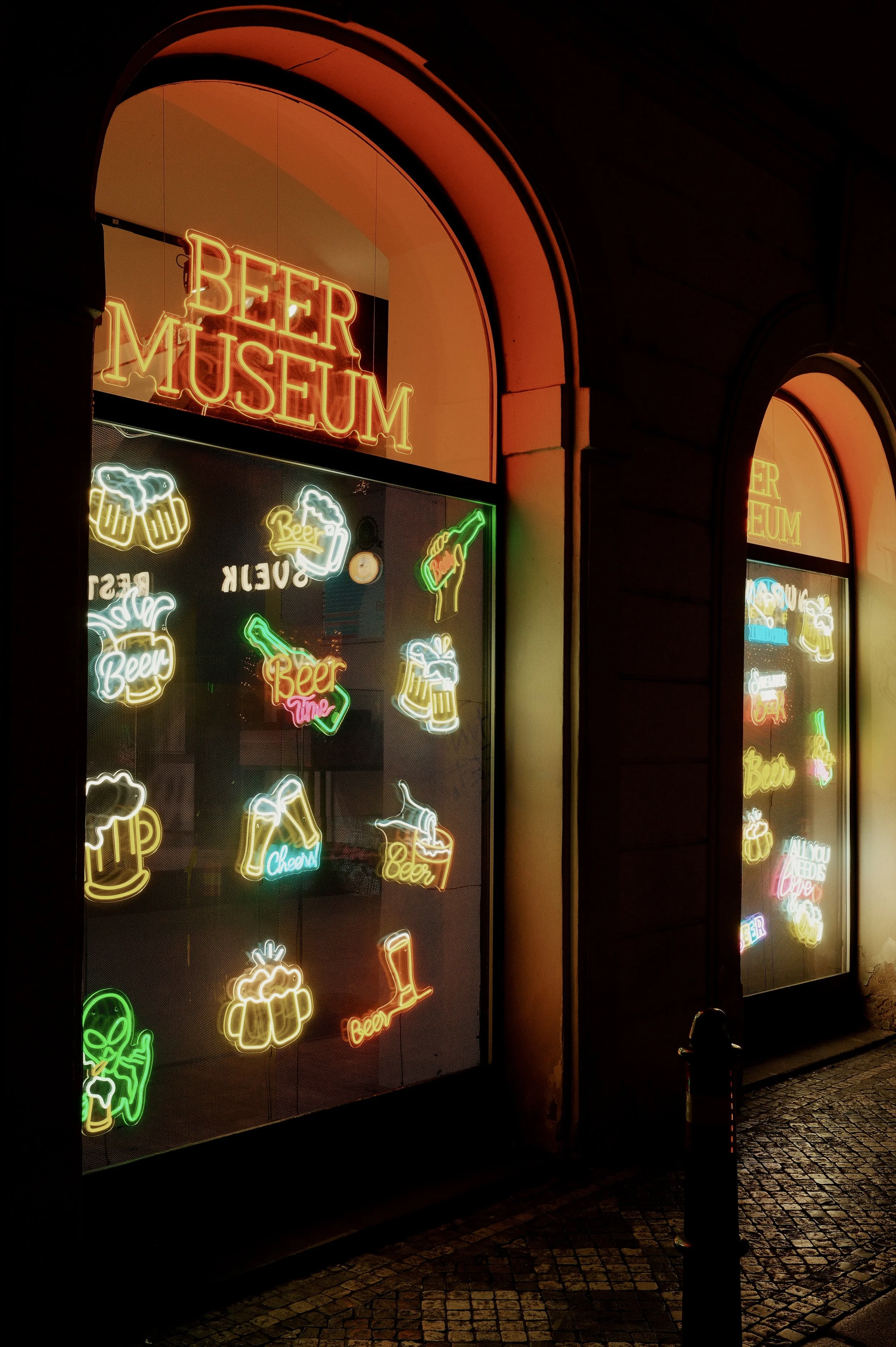 Night view of a beer museum storefront illuminated with colorful neon signs of beer mugs, bottles, and cheers messages.