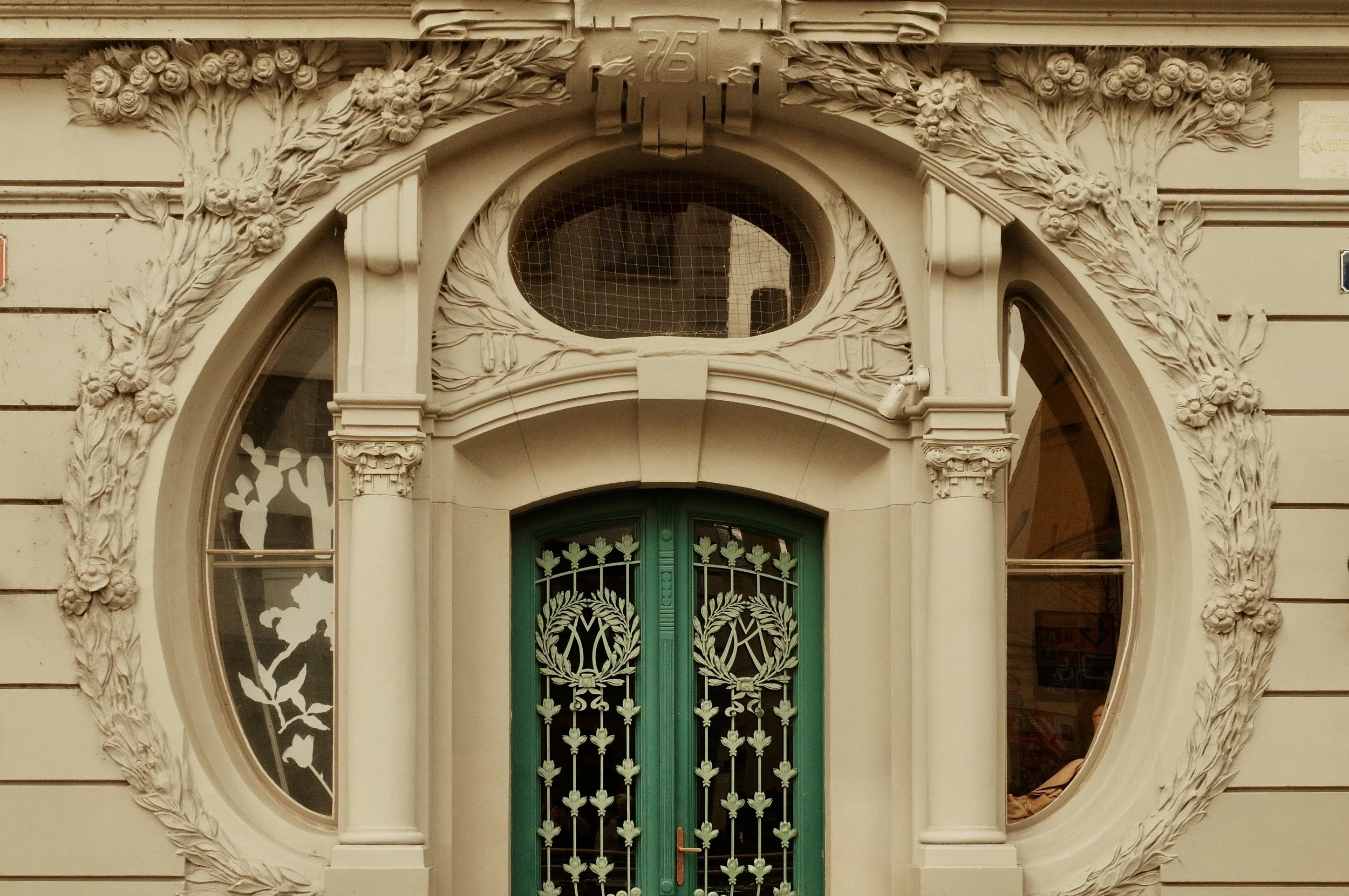 Ornate building facade featuring a central green door with decorative metalwork, oval and arched windows on either side, all surrounded by detailed floral and scroll ornamentation.