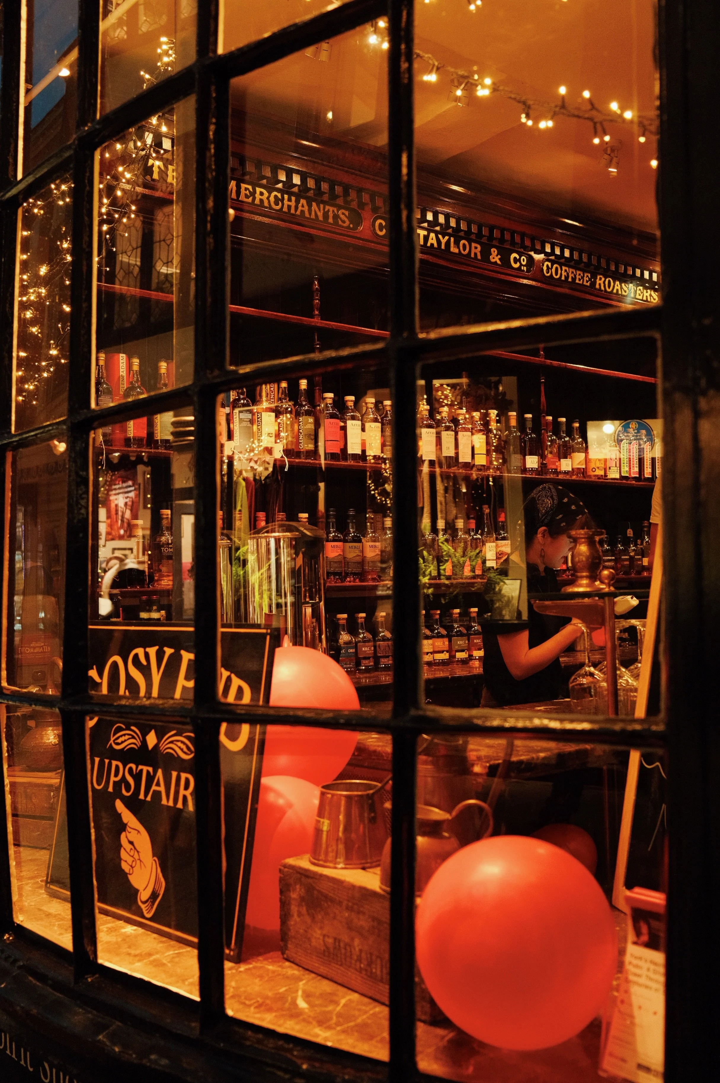 A cozy bar scene viewed through a window with black grid frames. Inside, shelves are stocked with numerous bottles of liquor, and warm ambient lighting with string lights creates a festive atmosphere. There are peach-colored balloons in the foregroun