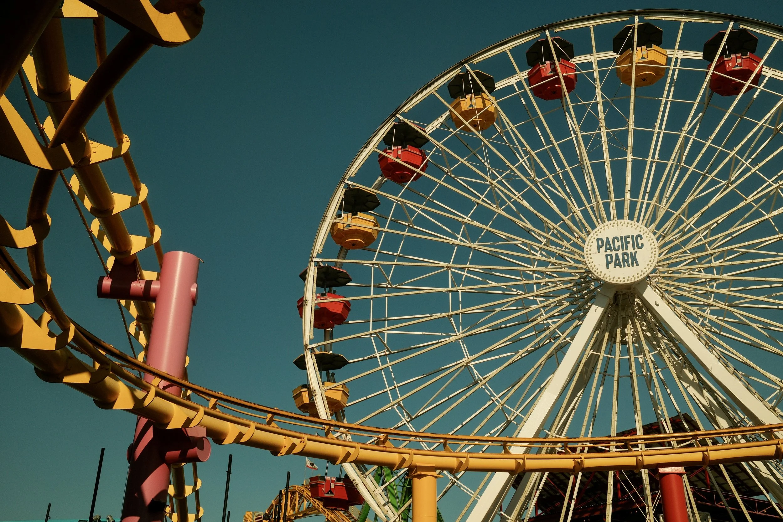A large Ferris wheel at Pacific Park with colorful gondolas against a clear blue sky, and part of a yellow roller coaster in the foreground.