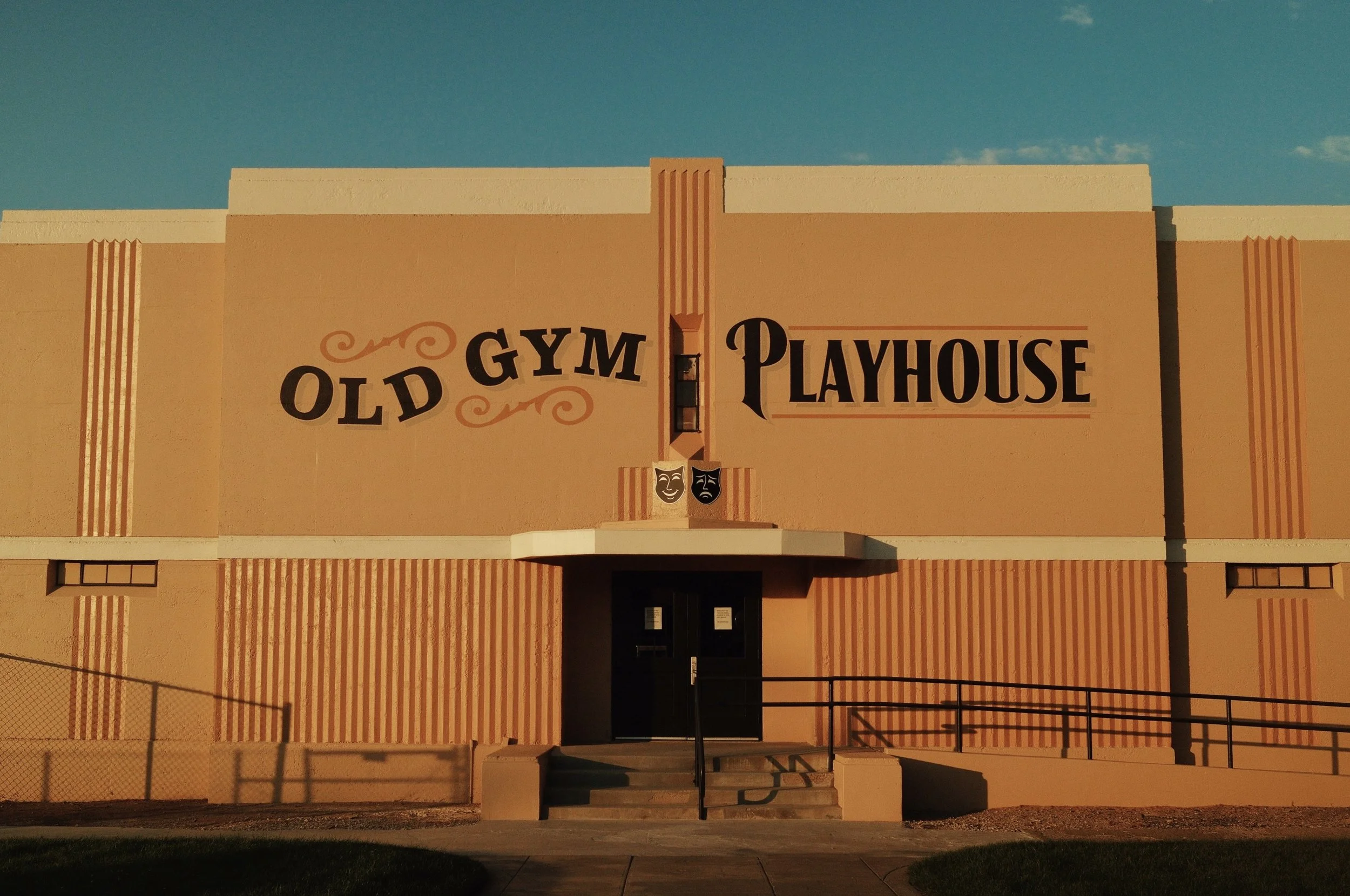 The front of a beige building with the sign 'Old Gym Playhouse' in black and brown lettering, featuring two theatrical masks above the entrance, a set of stairs, and a blue sky background.