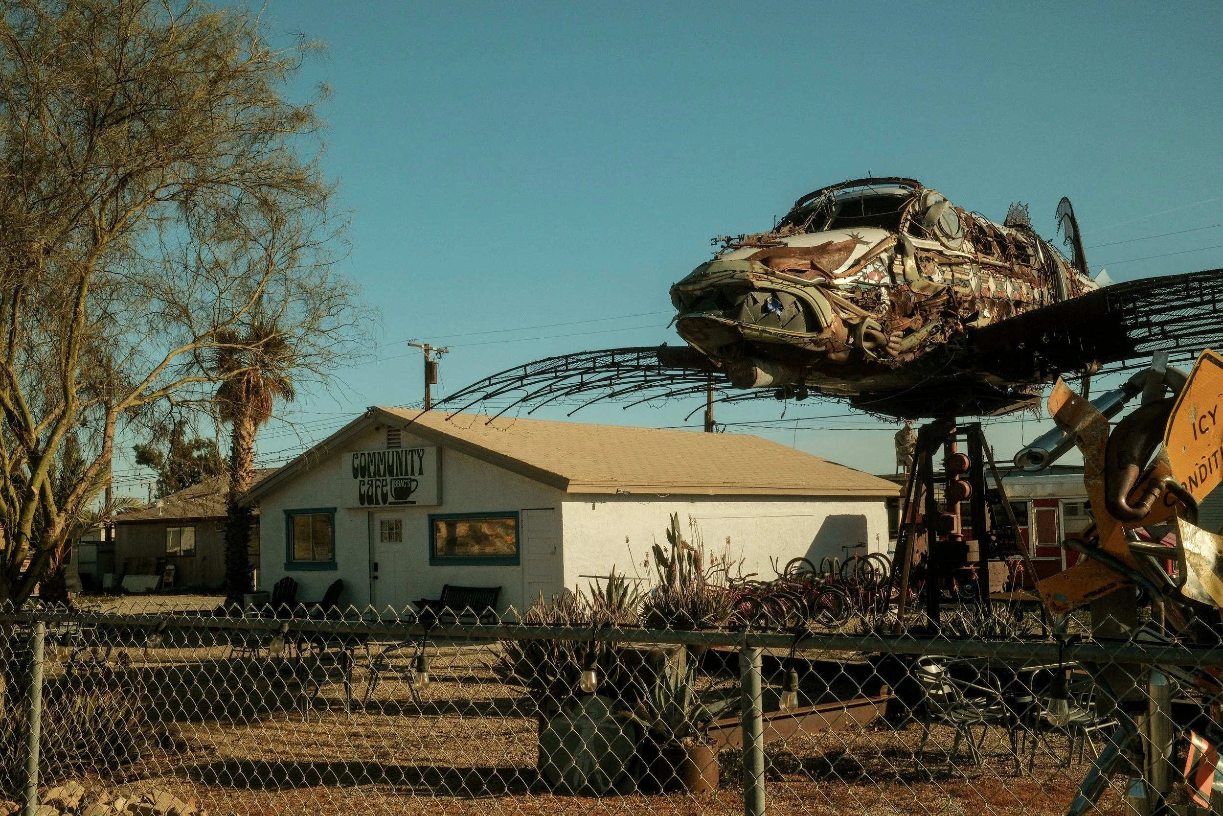 A large metal sculpture resembling a helicopter, made from various scrap metal pieces, is mounted on a platform in front of a white building labeled 'Community Cafe' in a small yard with a chain-link fence, cacti, and bikes nearby.