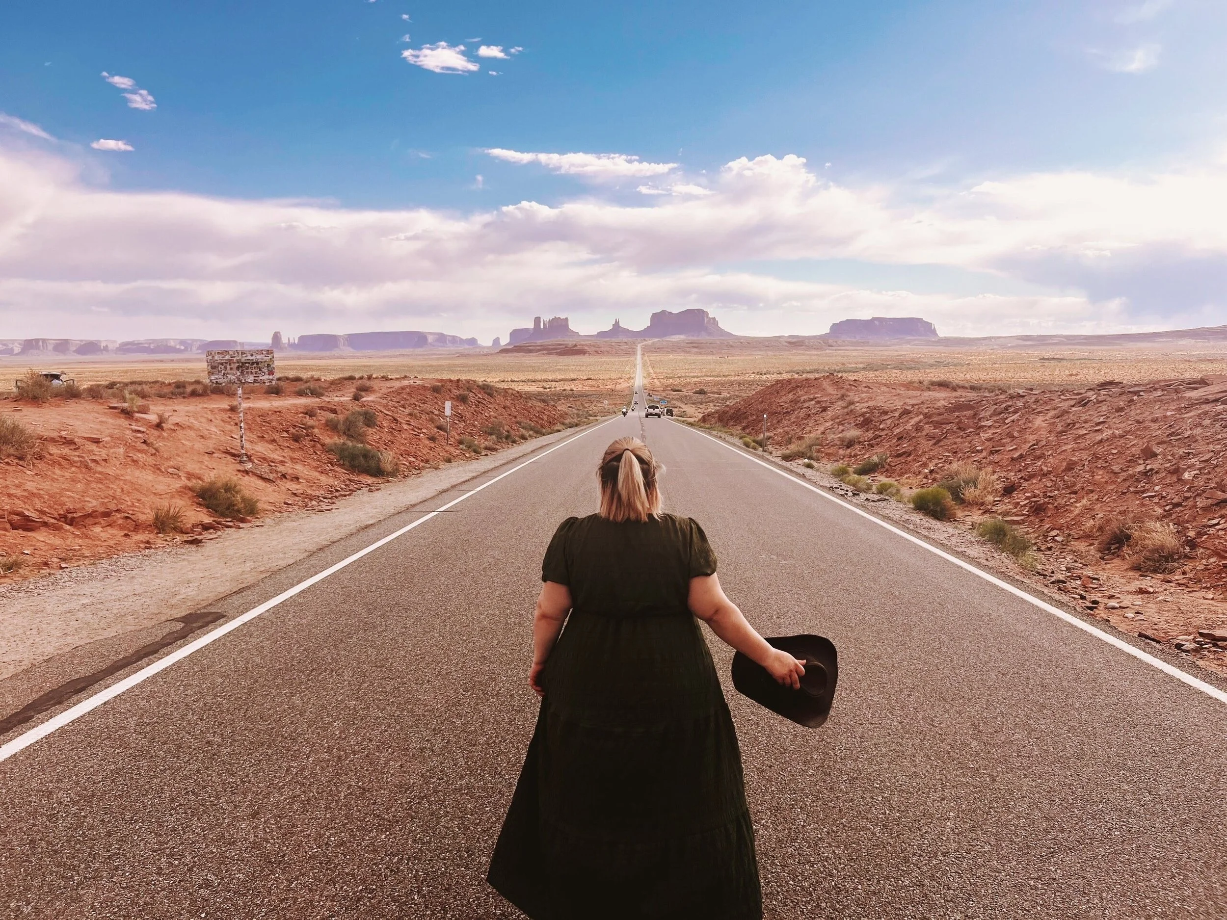 Woman with blonde hair holding a cowboy hat, standing in the middle of a long desert highway with red rock formations and blue sky in the background.