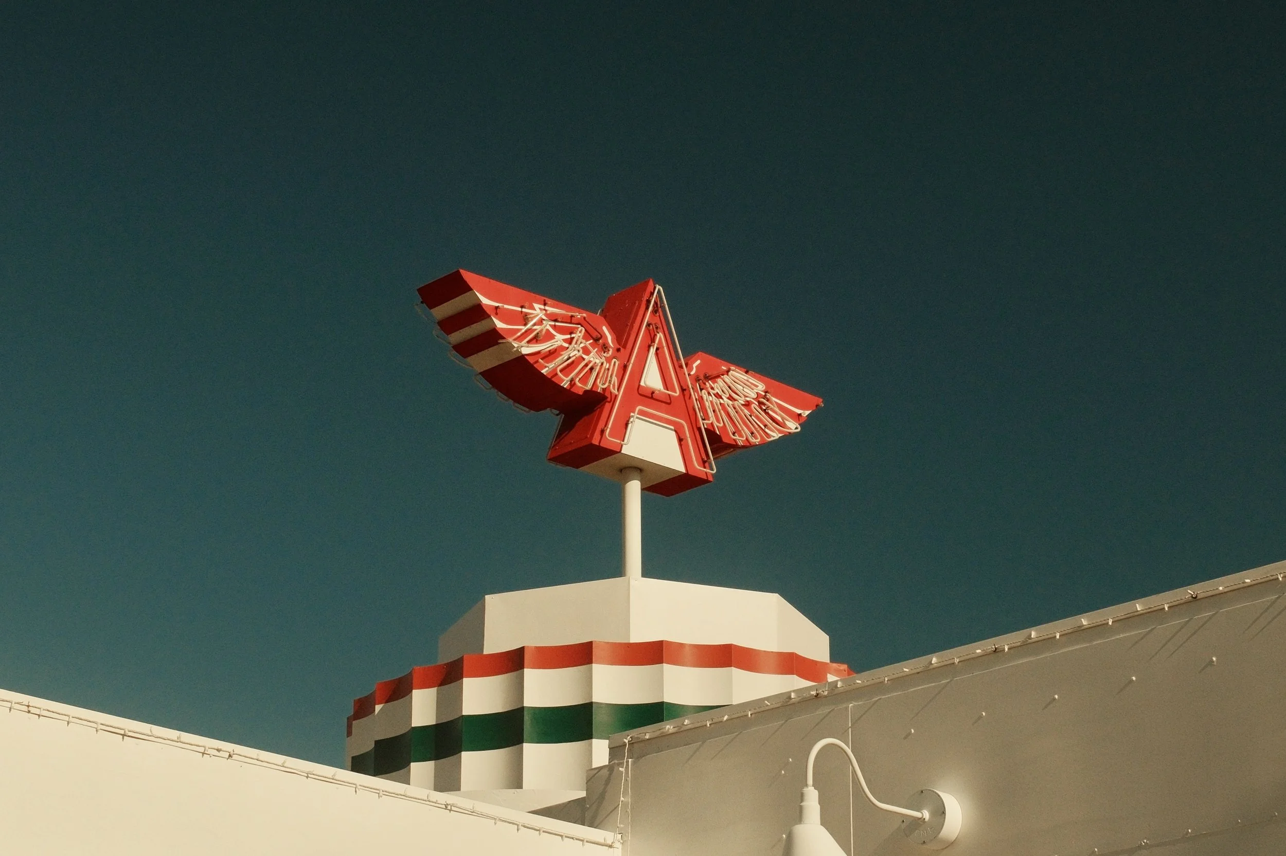 Retro neon sign with a red letter 'A' with white wings, mounted on top of a building with red, white, and green stripes, against a dark sky.