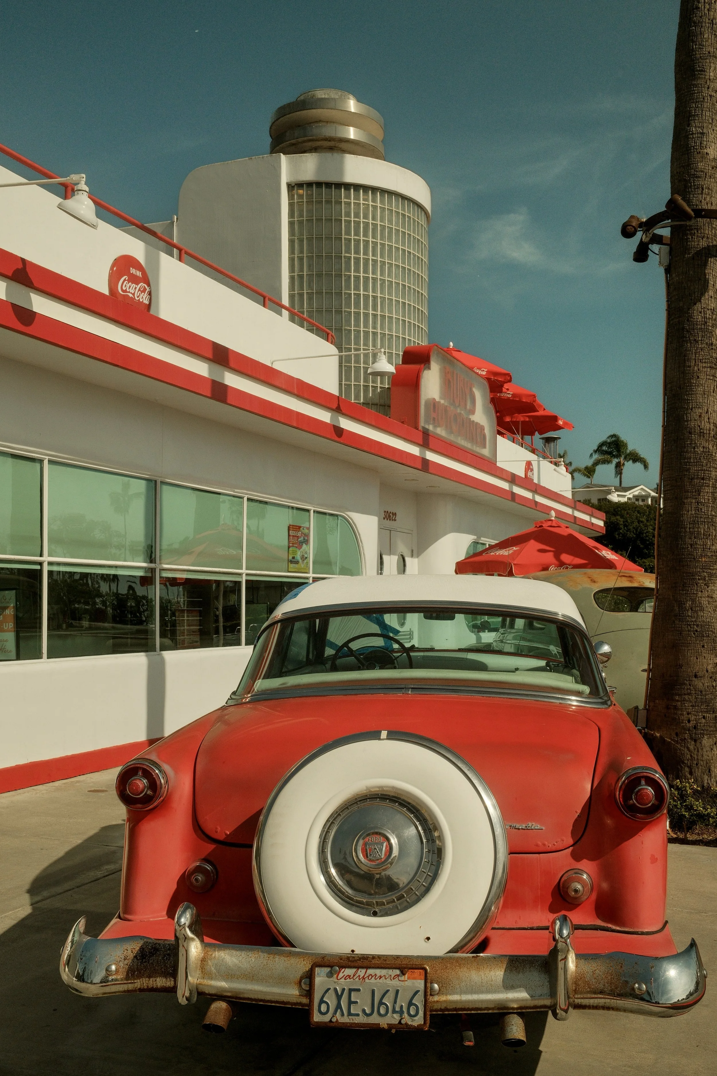 A vintage red and white car parked outside a classic diner with a Coca-Cola sign on the building and a tall building with glass blocks in the background.