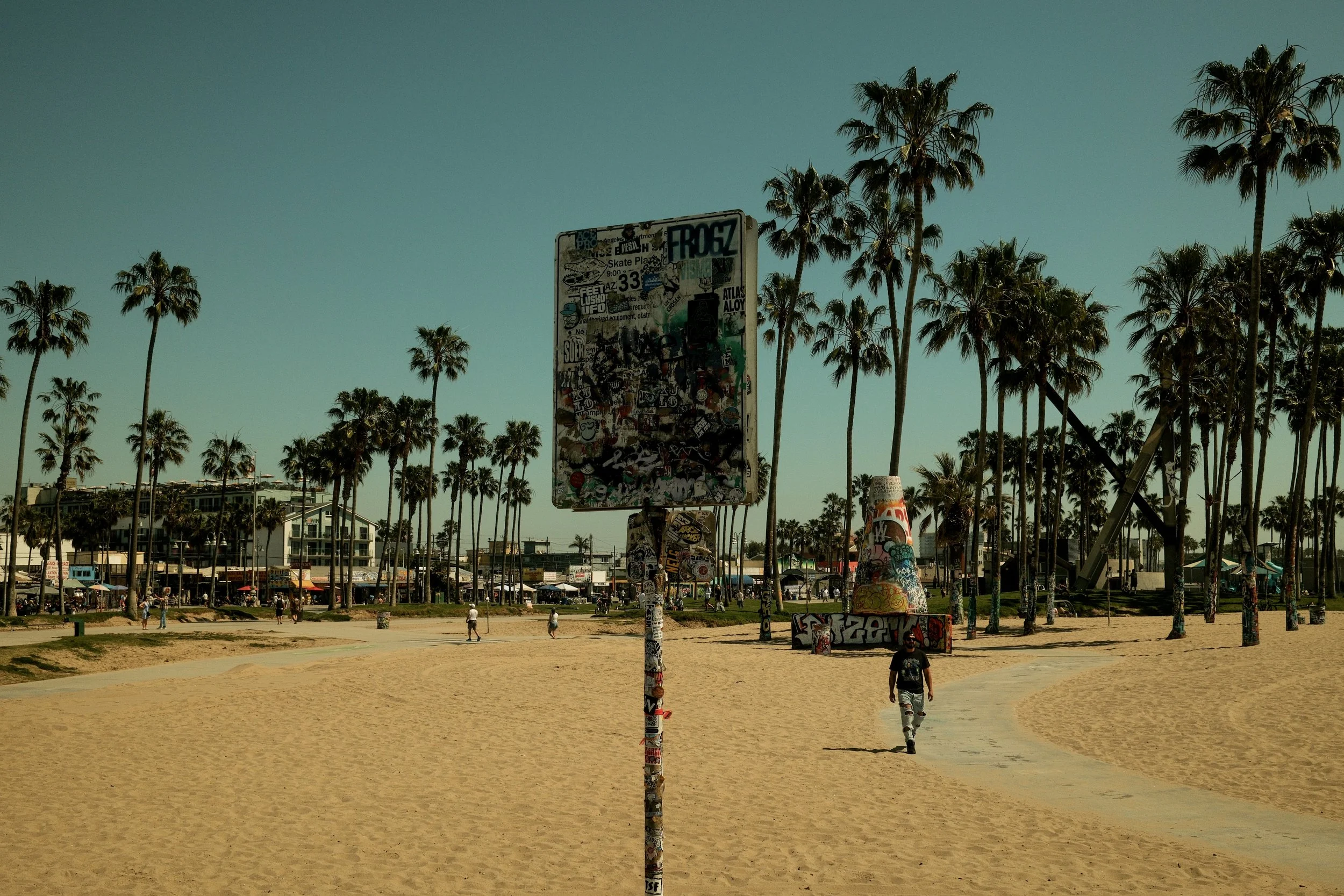 A sandy beach park with tall palm trees, graffiti-covered pole, pathway, and a person walking, with buildings and people in the background under a clear blue sky.