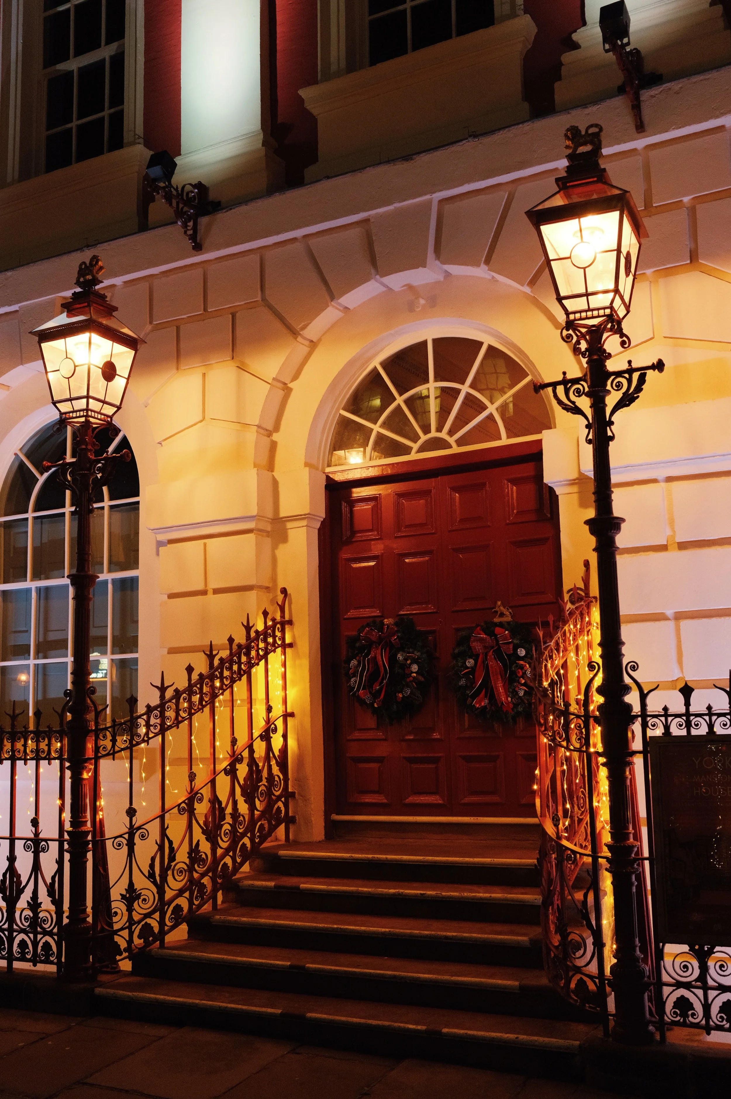 Decorative entrance of a building at night with two lit street lamps, a wooden door decorated with Christmas wreaths, and warm lighting.