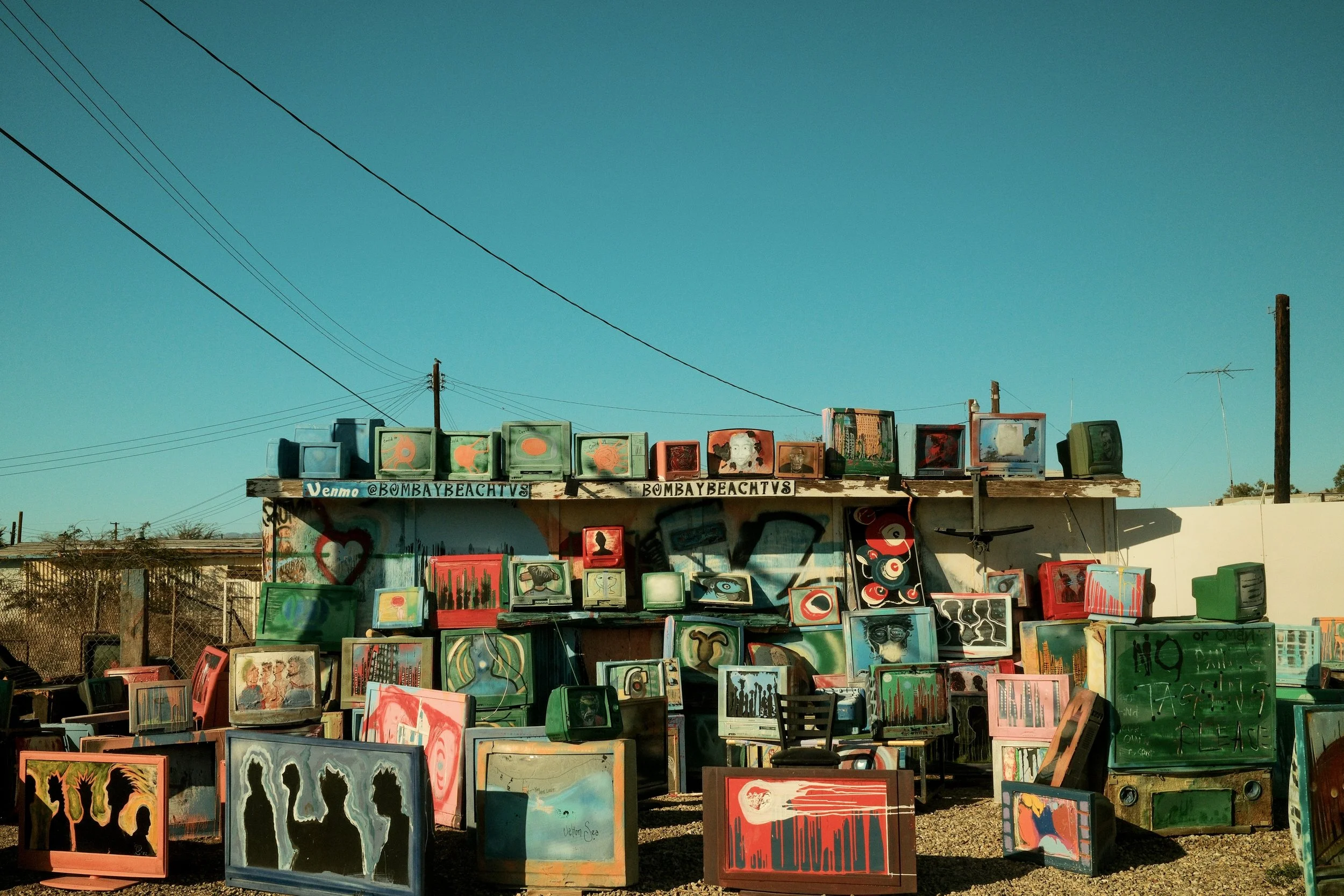 Outdoor art installation featuring multiple vintage televisions painted with abstract and colorful designs, arranged on shelves and on the ground under a clear blue sky.