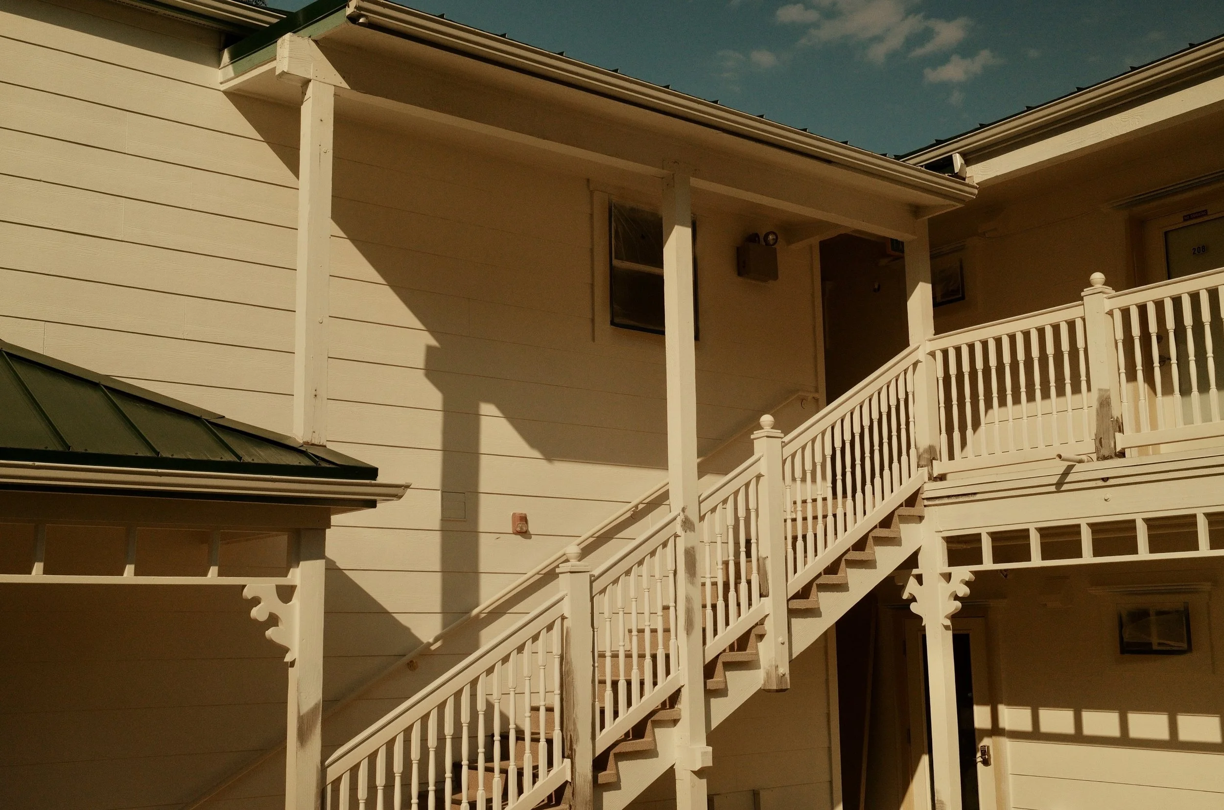 Close-up of a stairway leading to the entrance of a beige house with a balcony and a metal roof, under a blue sky with a few clouds.