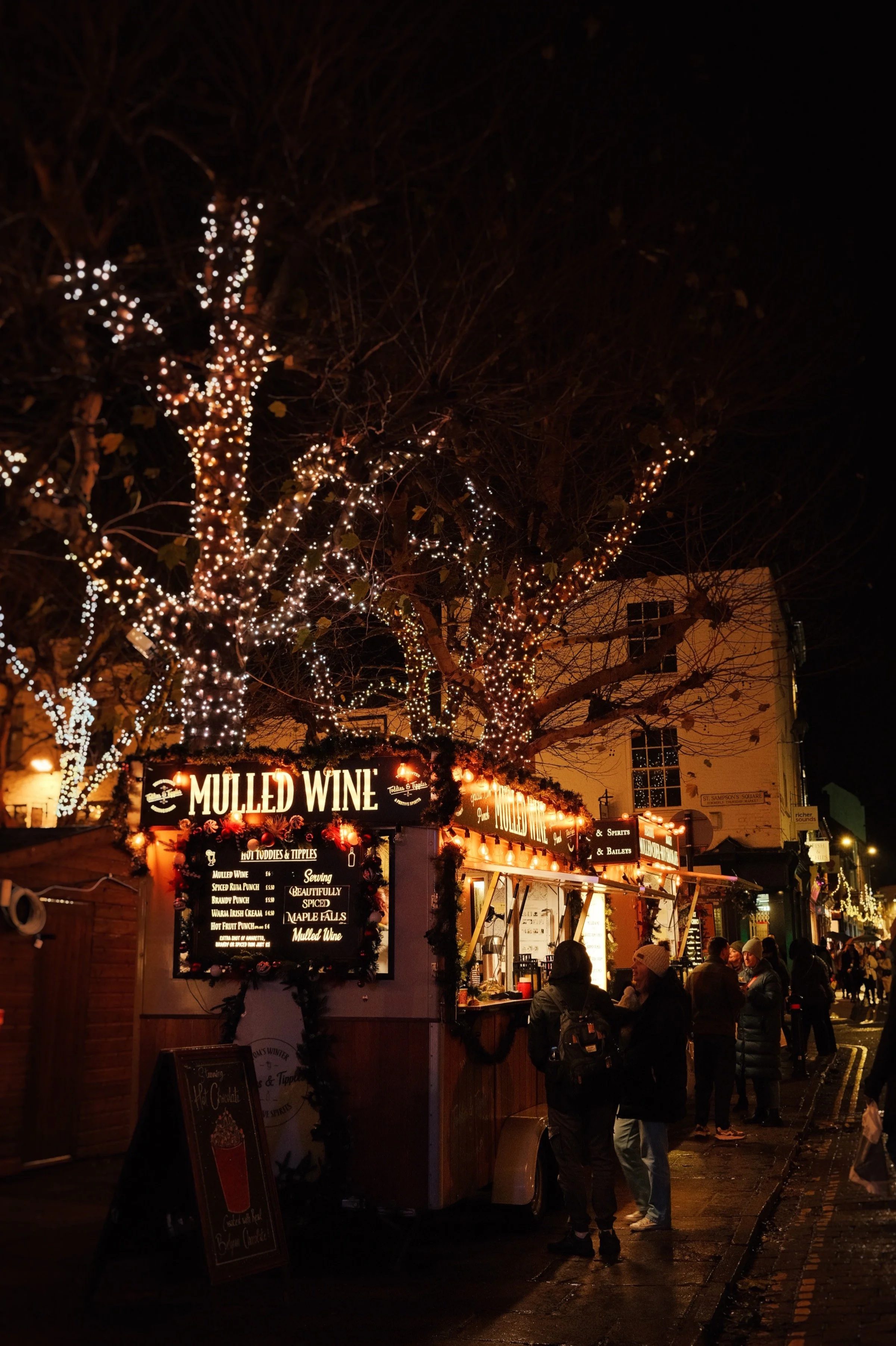 Night scene of a festive food stall decorated with lights, selling mulled wine, with people standing and chatting in front, and trees with twinkling lights overhead.