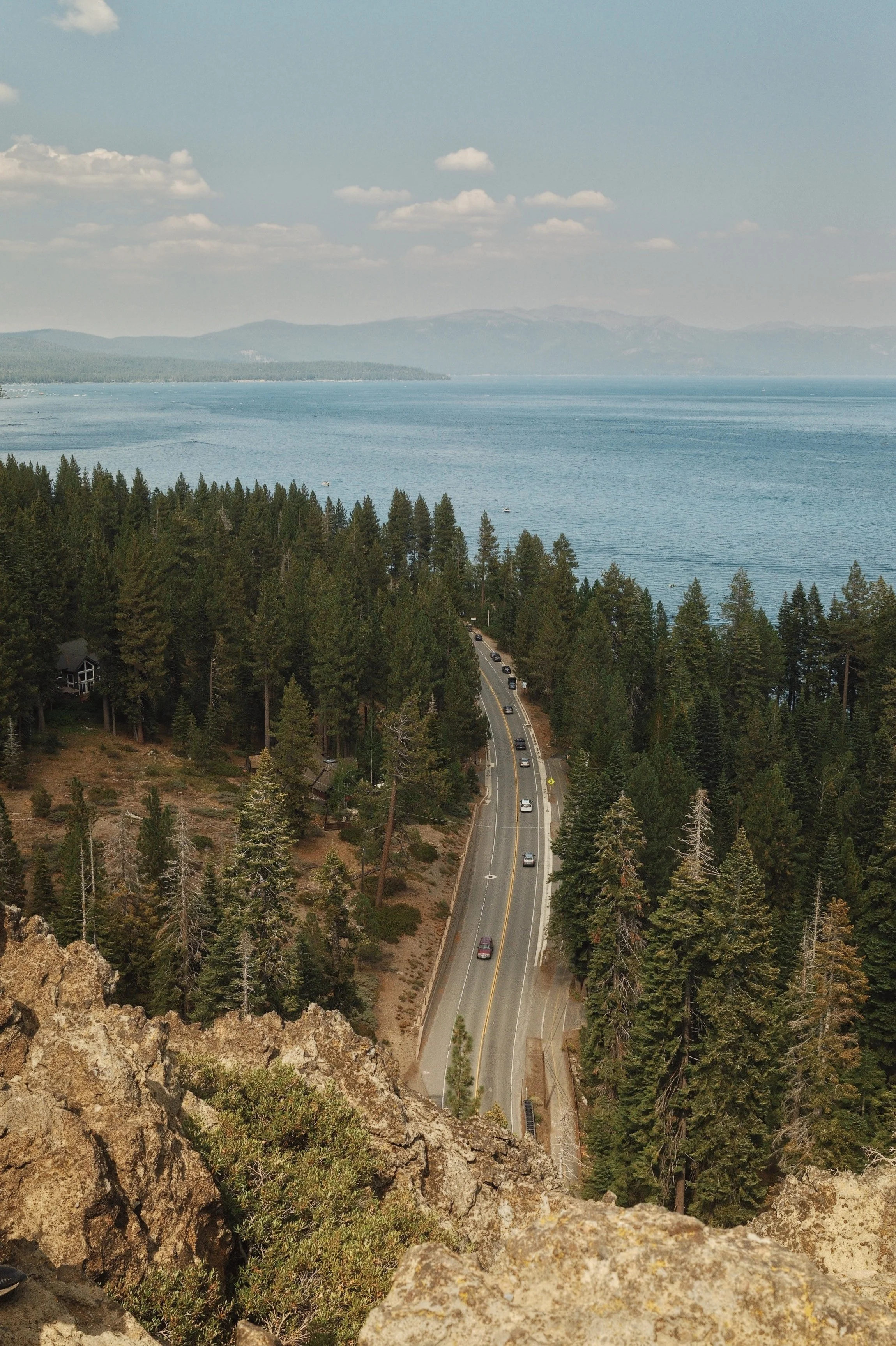 A scenic view of a winding road through a forested area beside a large body of water under a partly cloudy sky.