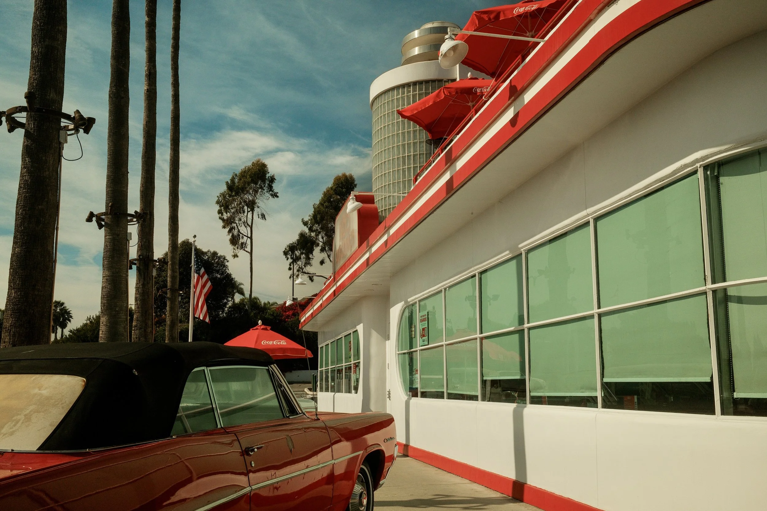 A vintage red convertible car parked outside a retro-style diner with large windows, green curtains, red umbrellas, and an American flag in the background, under a partly cloudy sky.