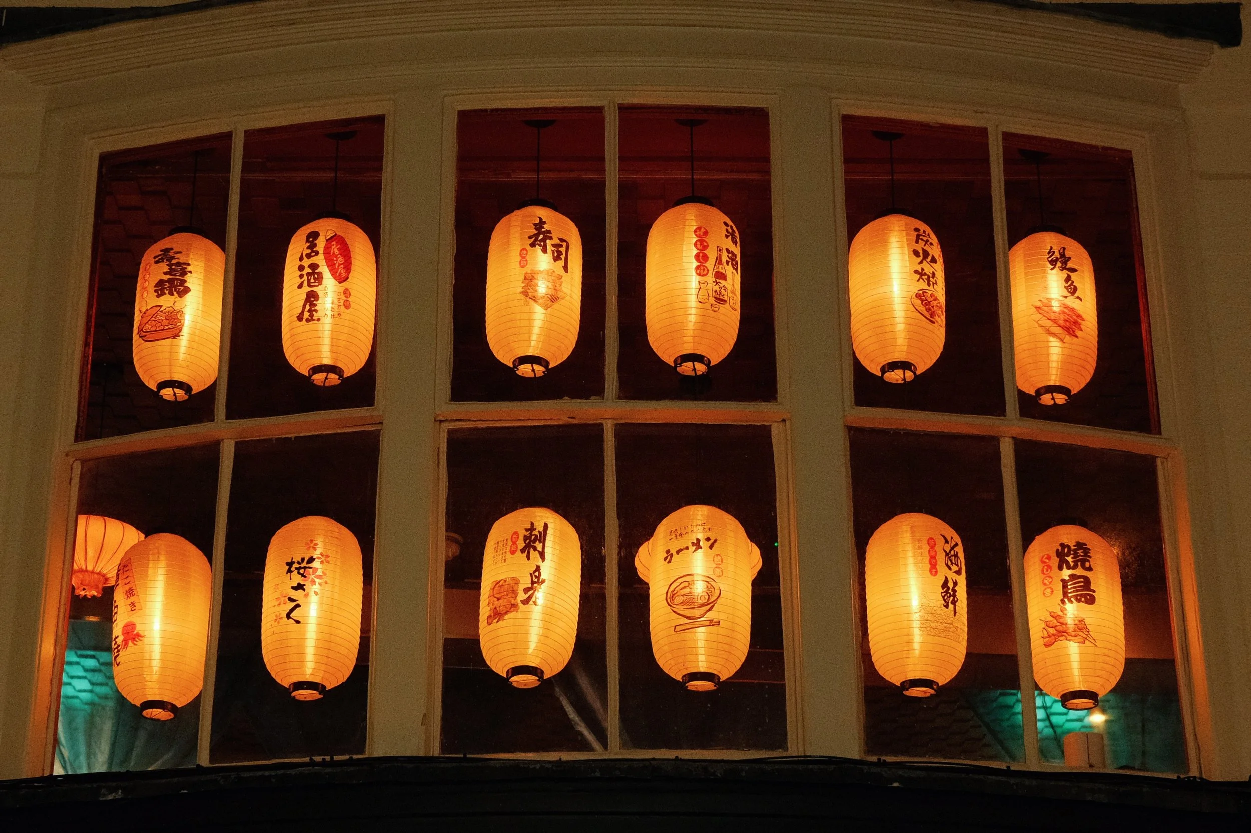 Multiple orange Japanese paper lanterns with black and red writing hanging in a window.