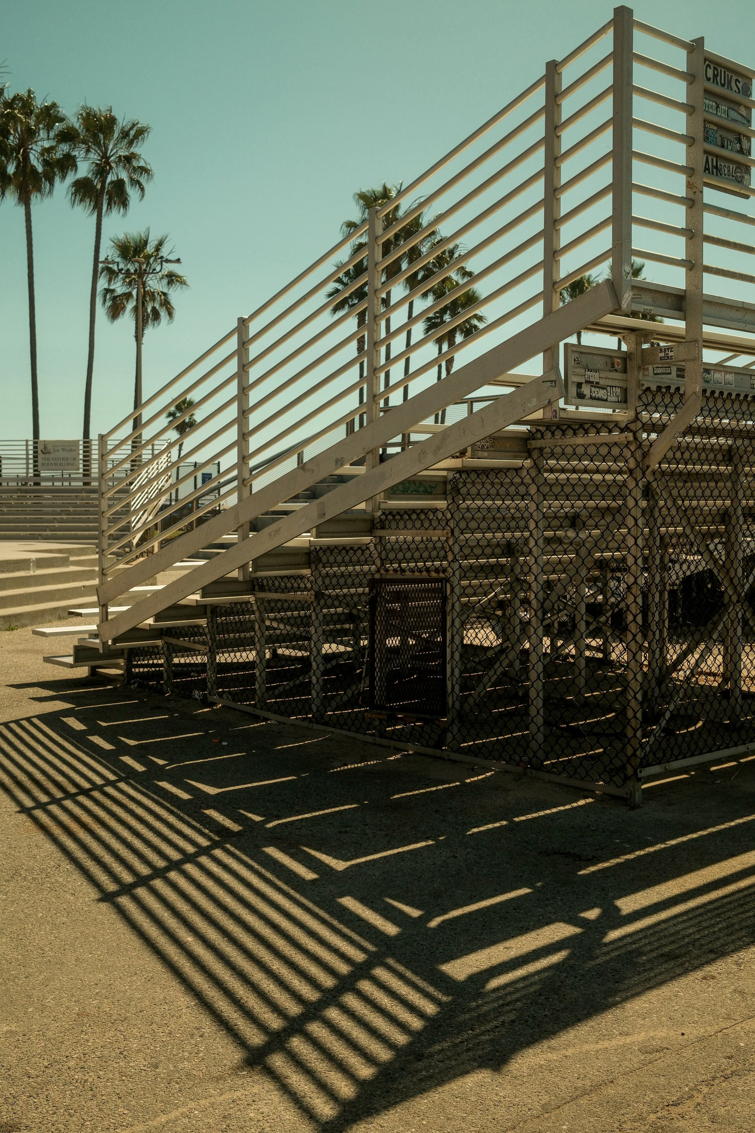 Empty outdoor bleachers with palm trees in the background, casting shadows on the ground under a clear sky.