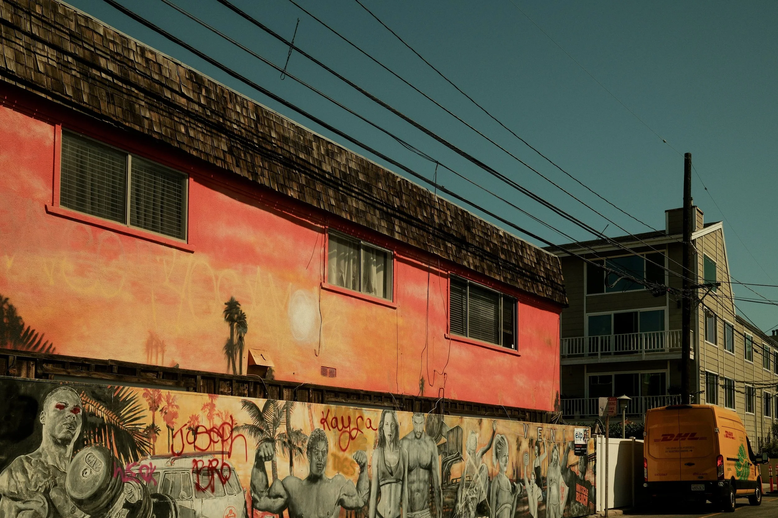 Street view showing two buildings, one with a pink and orange mural and graffiti, and the other with beige siding. Graffiti features muscular figures and palm trees. A yellow DHL van is parked in front, and power lines cross the sky.