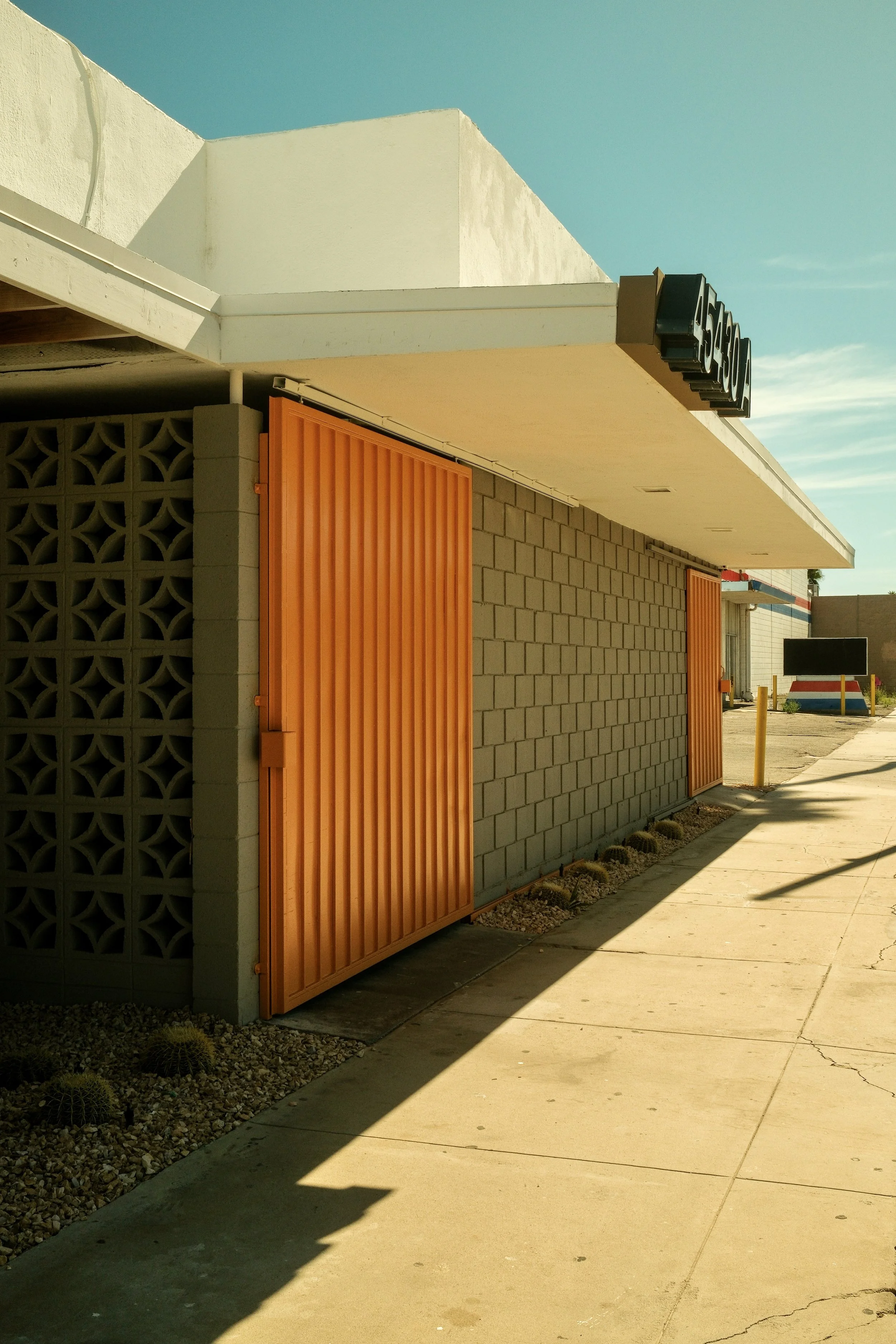 Side of a building with orange sliding doors, beige concrete block wall, and a white overhang, adjacent to a sidewalk with shadows.