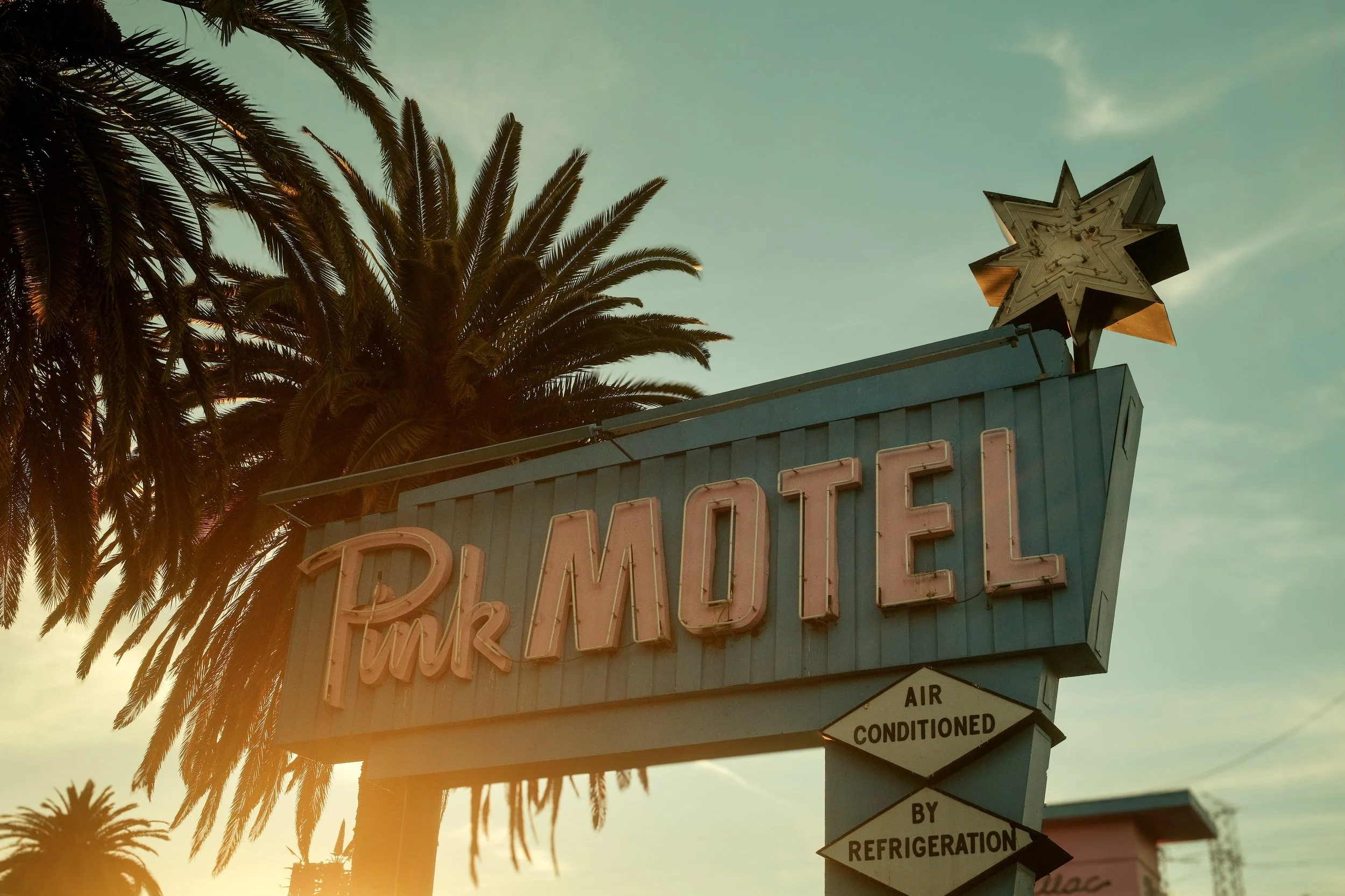 Vintage motel sign with palm trees in the background, featuring the word 'MOTEL' in large pink neon letters and the words 'Pink Motel' in smaller script. Additional signs indicate air conditioning and refrigeration.