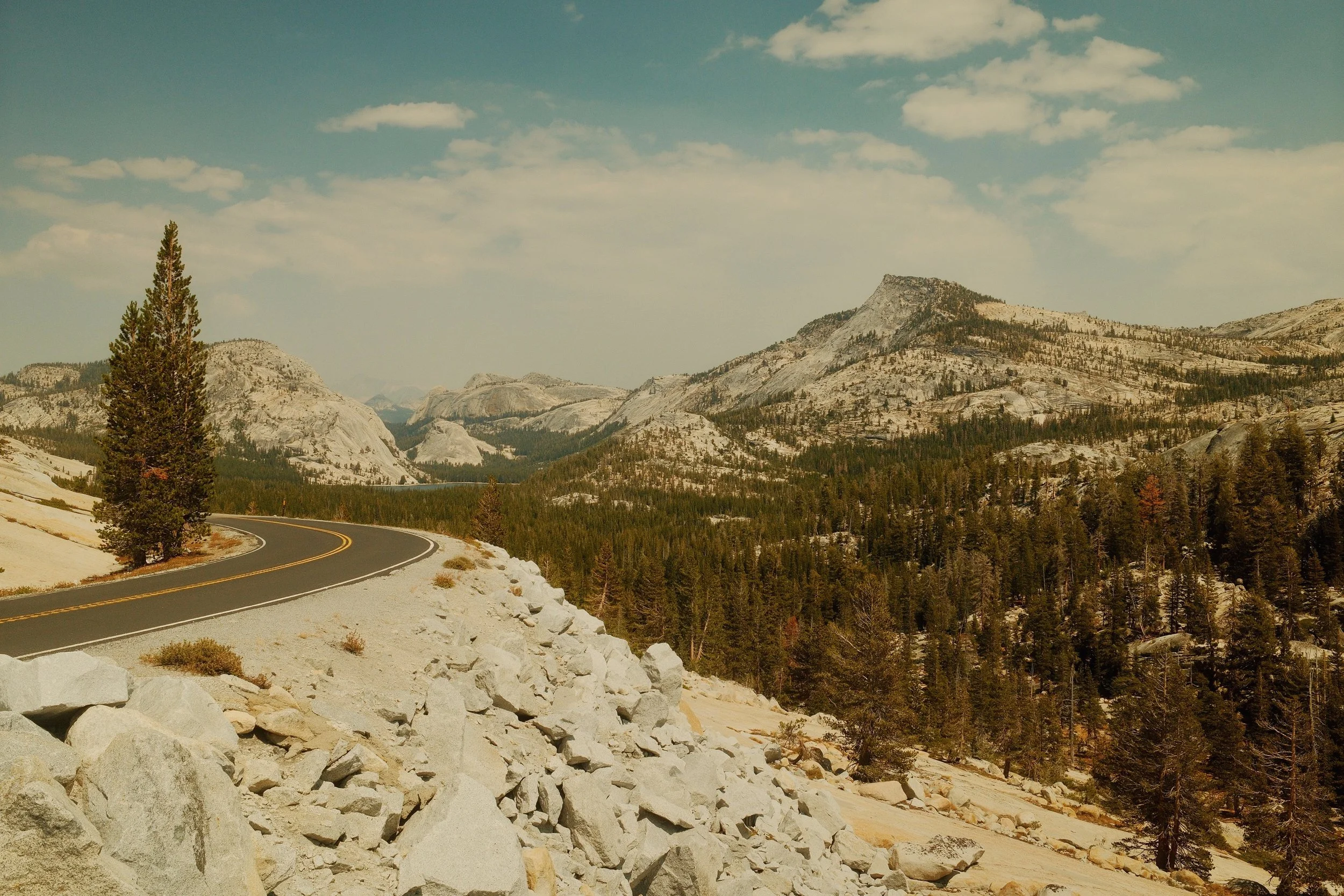 Scenic view of a winding mountain road through a forested valley with rocky hills and mountains in the background under a partly cloudy sky.
