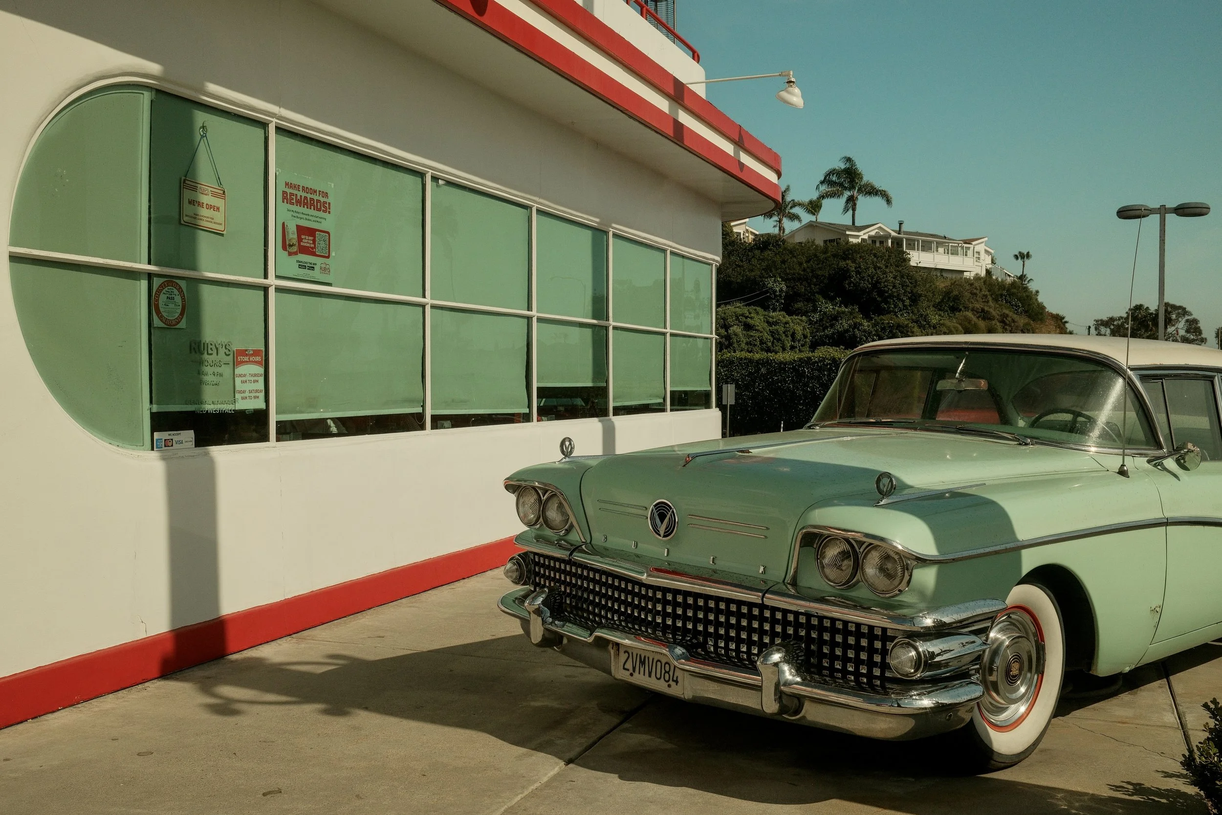 A vintage mint green car parked outside a retro-style diner with large glass windows and a red and white color scheme, in a sunny coastal town.