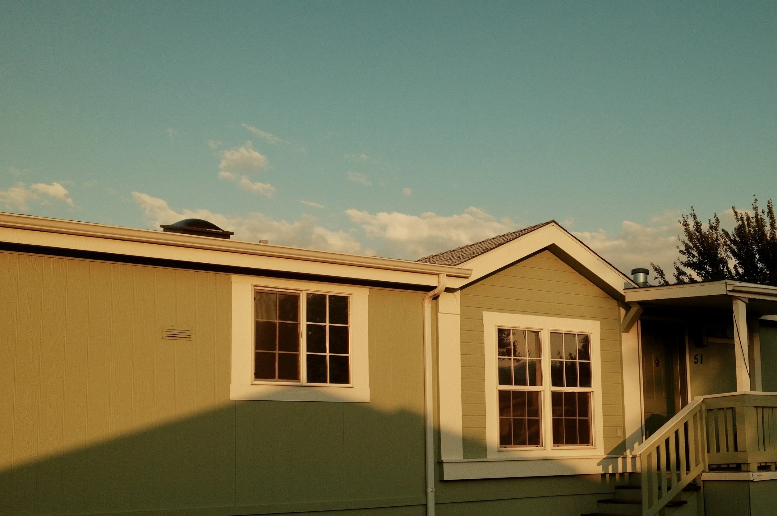 A house with green exterior walls, white window frames, and a staircase leading to the entrance, under a partly cloudy sky with sunlight.