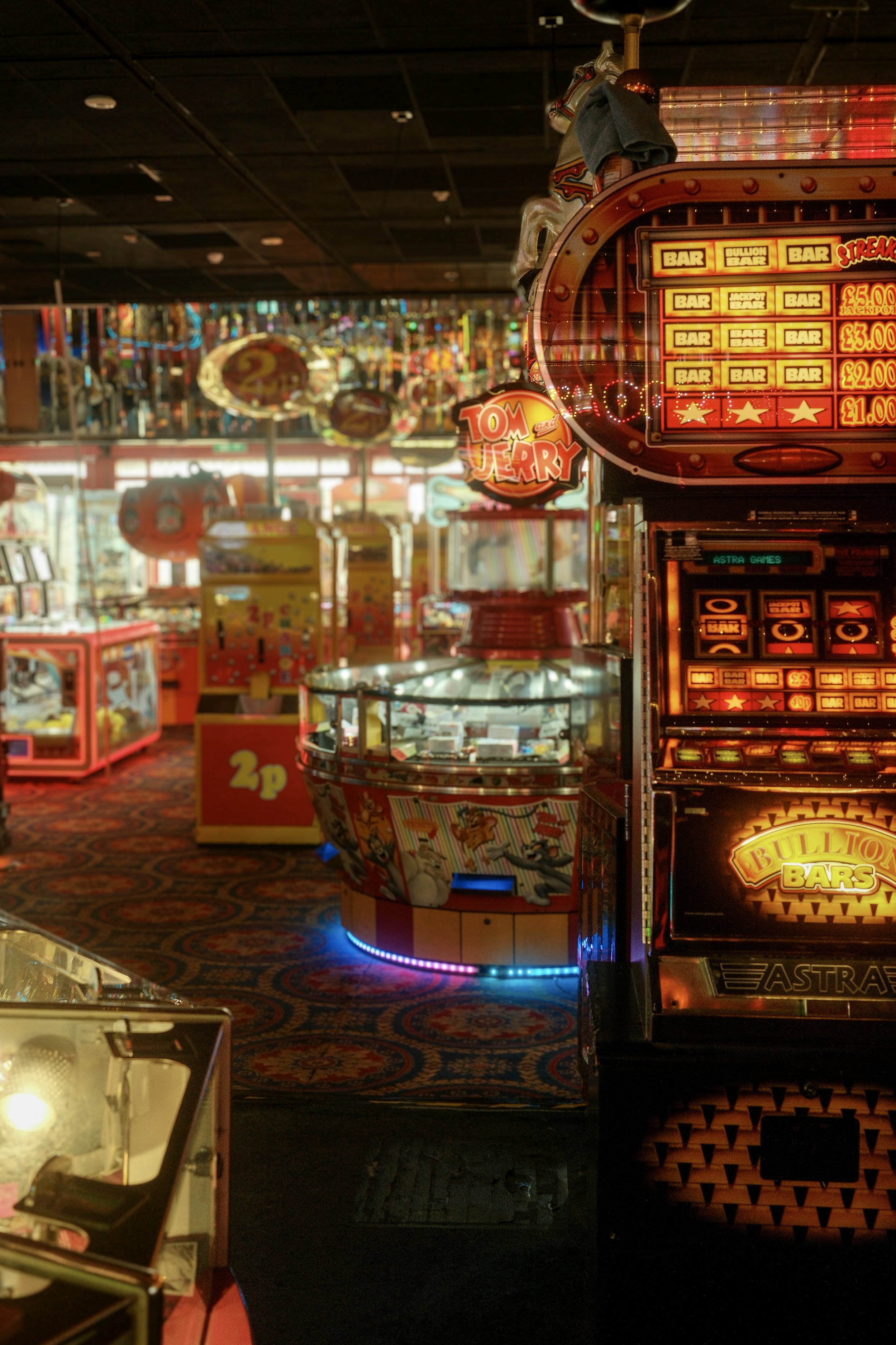 An arcade with electronic gambling machines, bright lights, and colorful decorations, featuring a slot machine and a carousel animal on the ceiling, with various game prize displays and a patterned carpet.