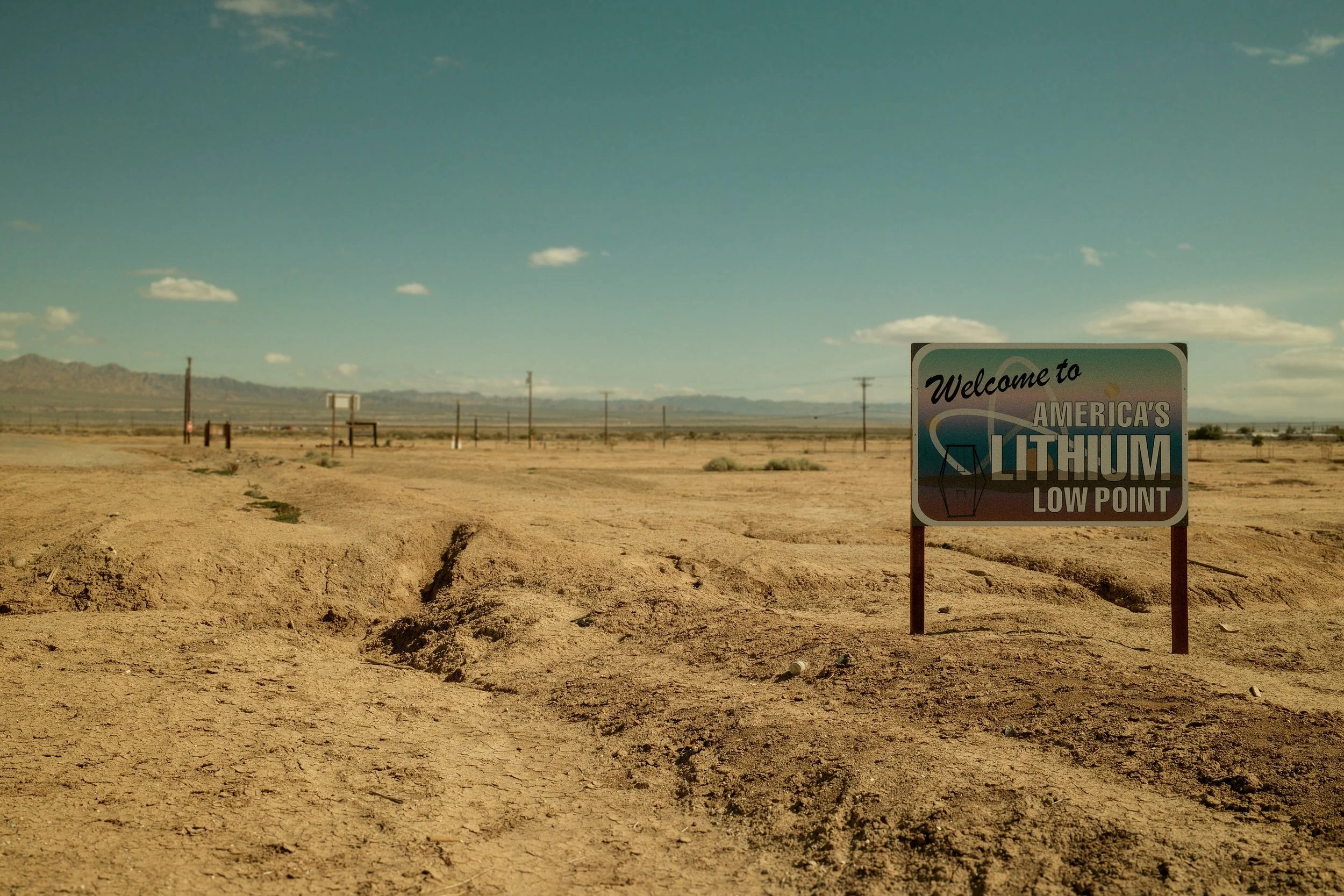 A sign reading 'Welcome to America's Lithium Low Point' in a barren desert landscape with mountains in the distance, clear blue sky, and utility poles.