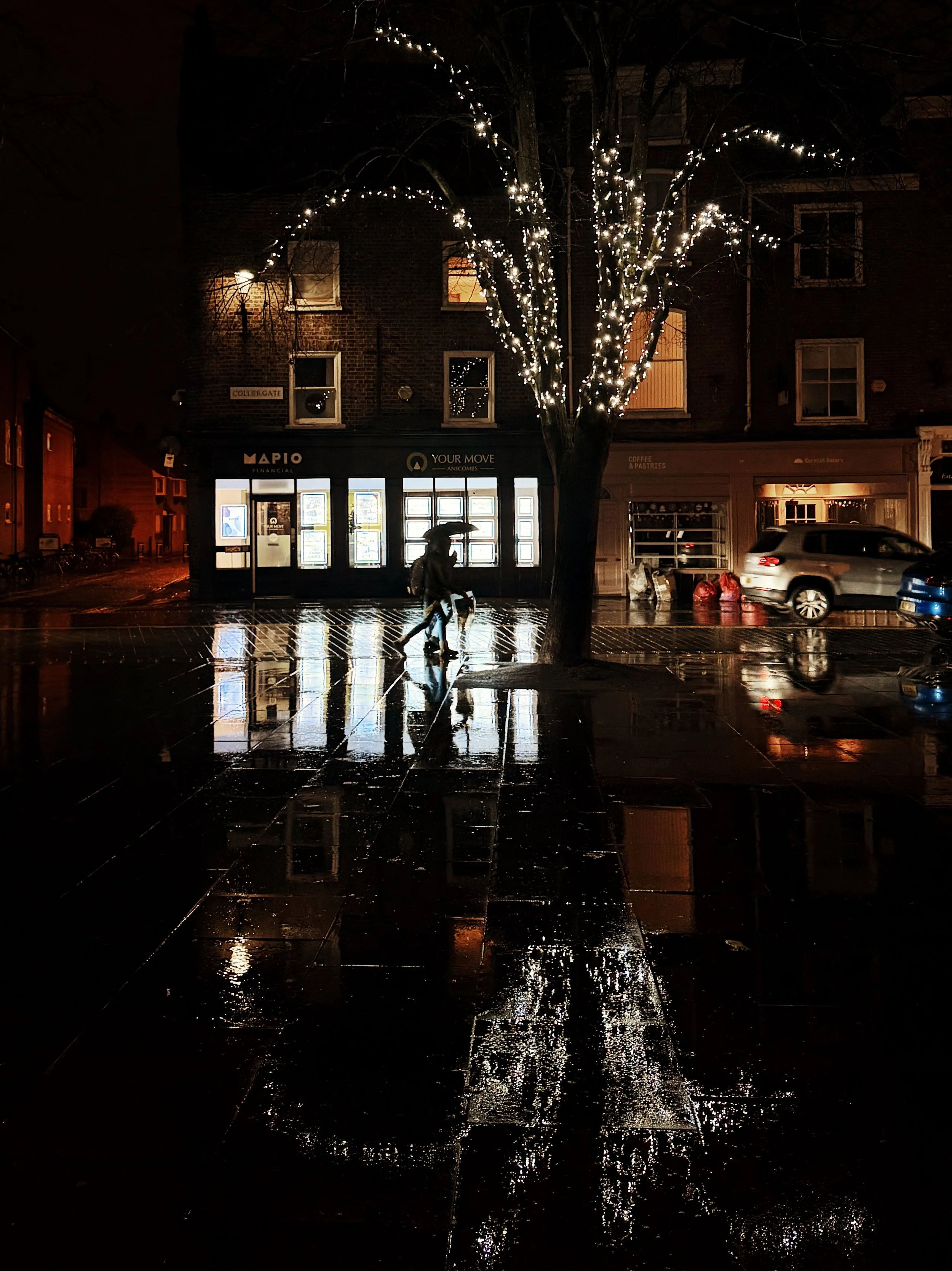 Nighttime city street scene with a lit tree, wet pavement reflecting lights, a person with an umbrella walking, parked cars, and illuminated storefronts.