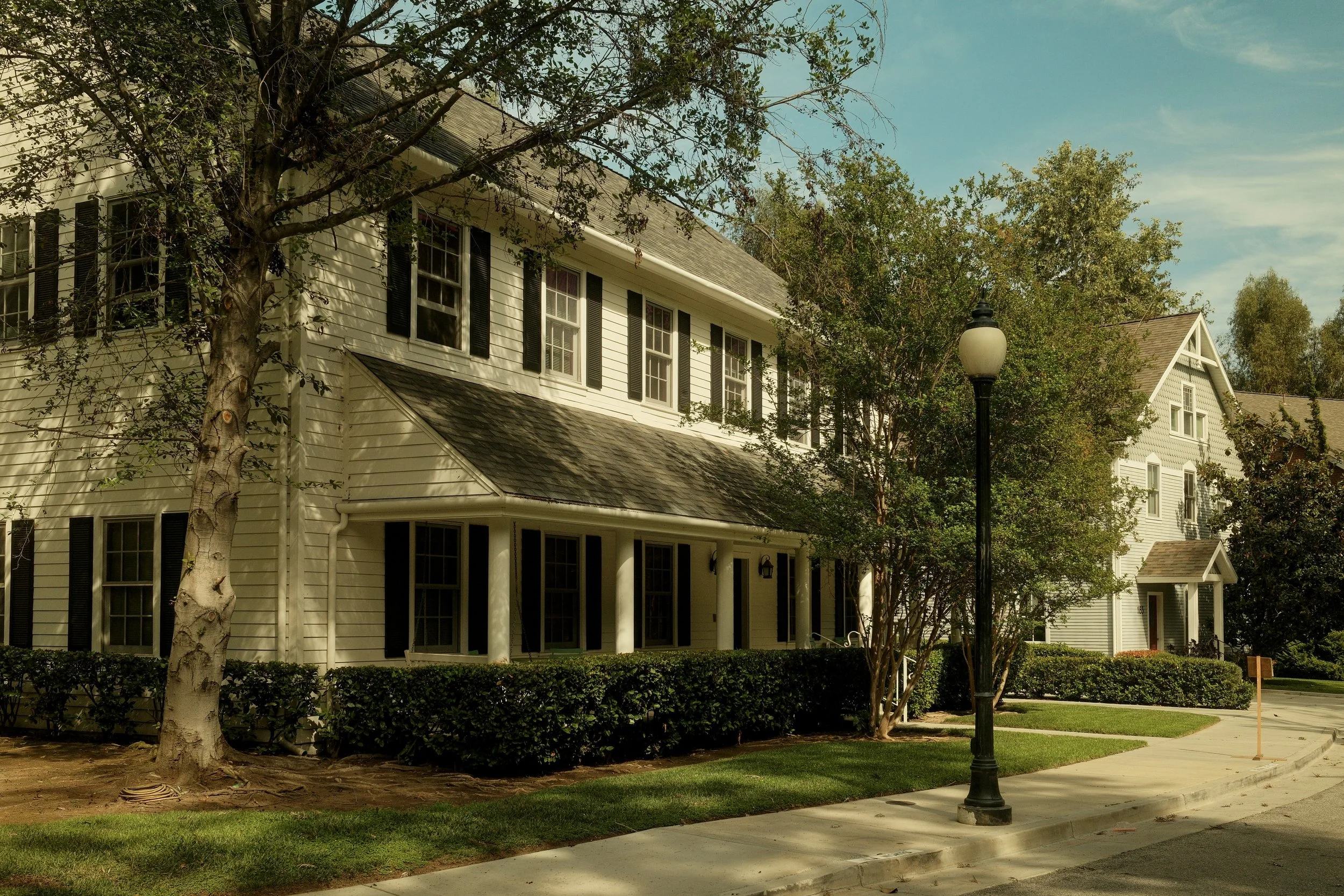 A row of white houses with black shutters on a sunny day, with well-maintained green lawns, trees, and a street lamp in the foreground.