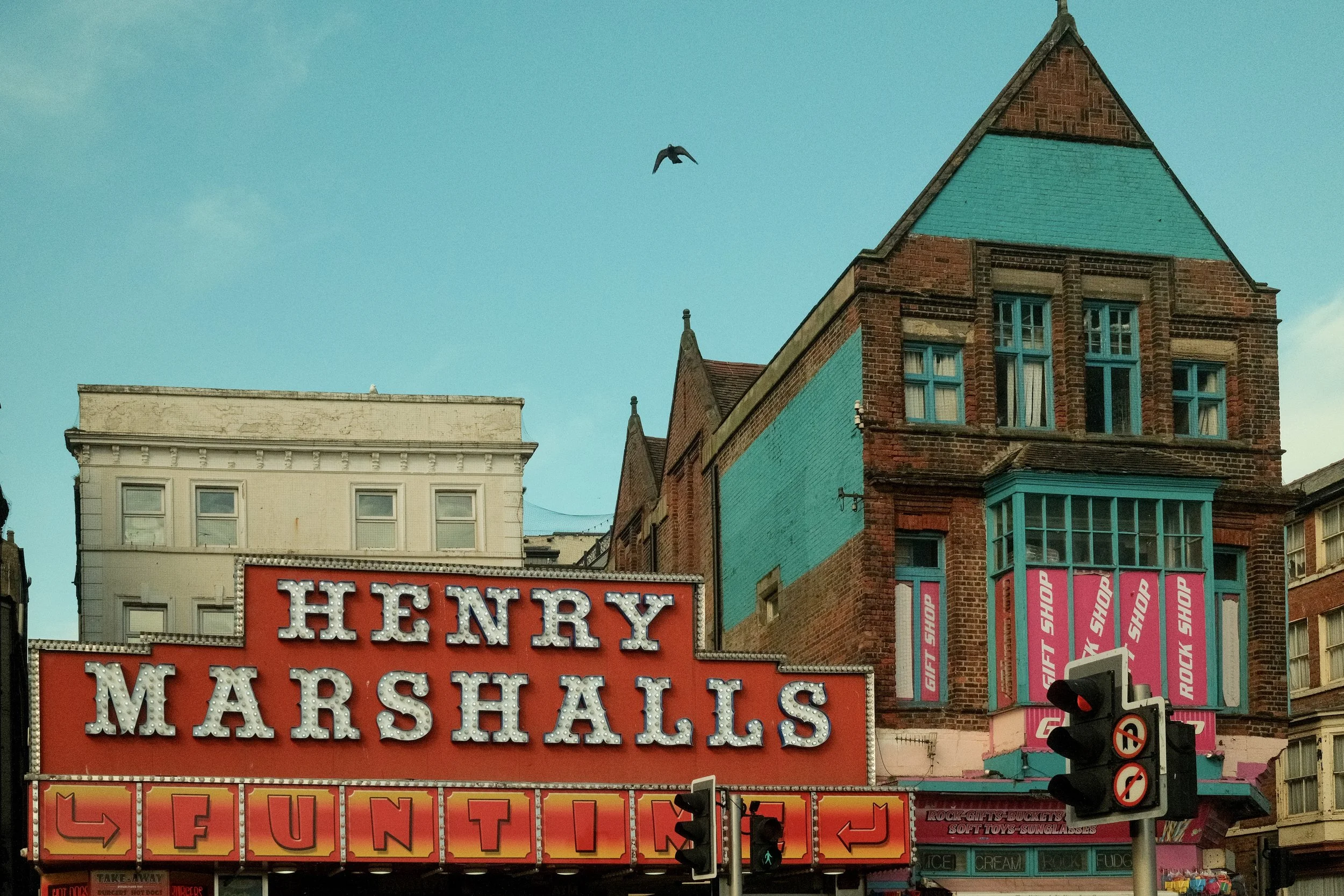 City street scene featuring a large red marquee sign for "Henry Marshalls" with bright bulbs, adjacent to a brick building with decorated windows, and a blue sky with a flying bird.
