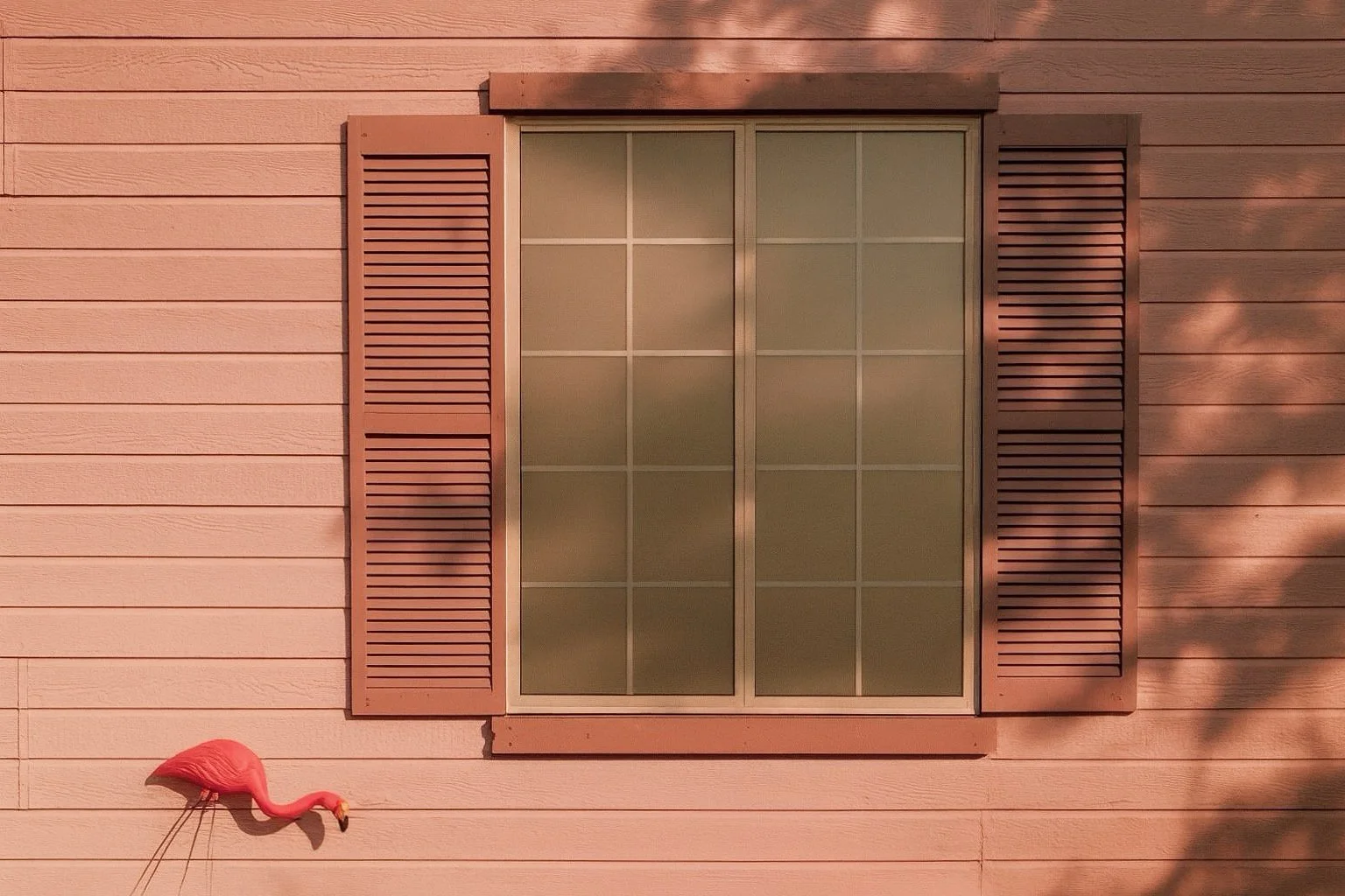 Pink house wall with horizontal siding, a window with beige shutters, and a decorative pink flamingo lawn ornament.