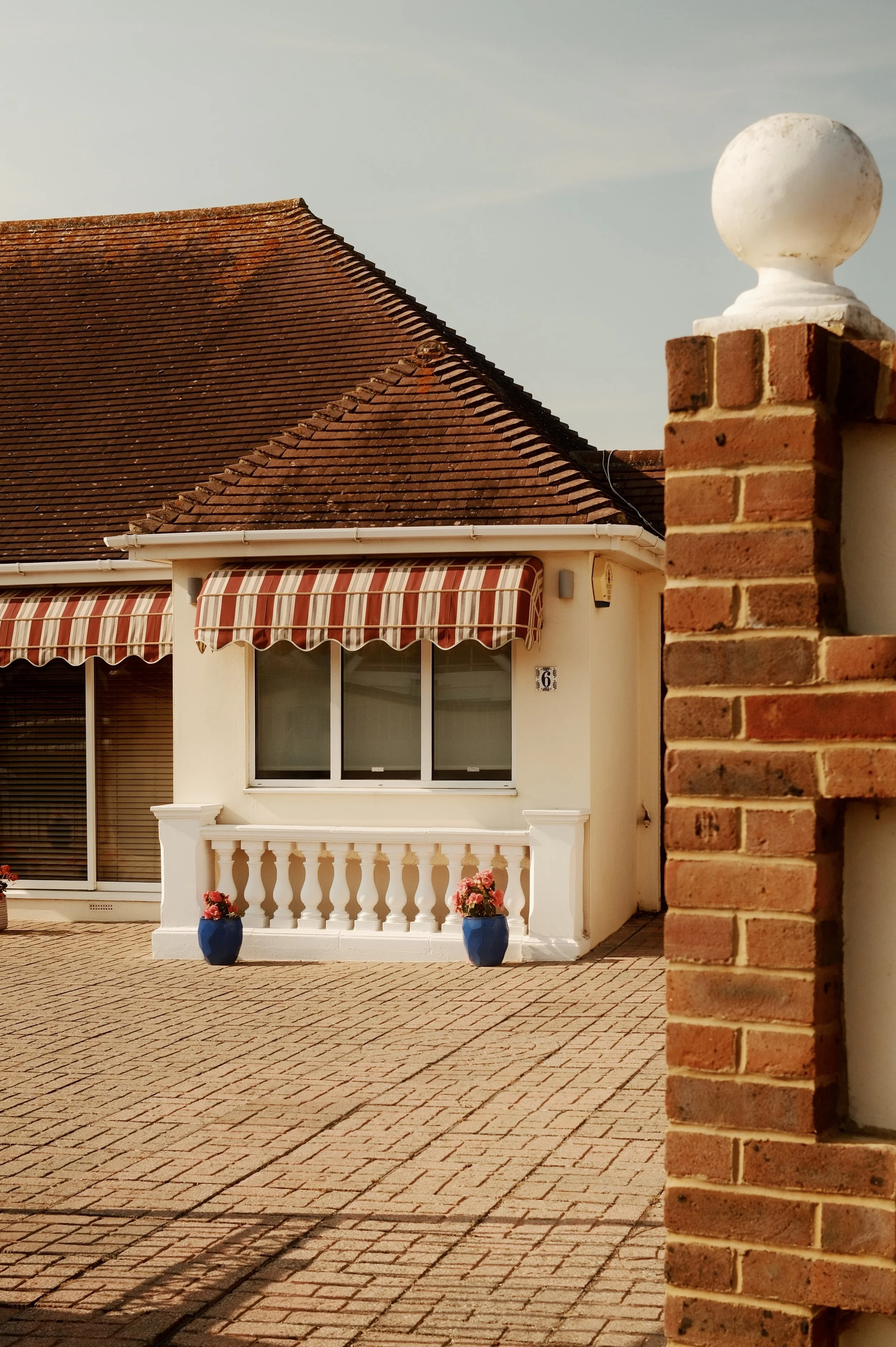 A house with a red tiled roof, white exterior walls, and a striped red, white, and beige awning over the window. There are two blue flower pots with pink flowers on the white porch railing. A brick wall with white ornament on top is in the foreground