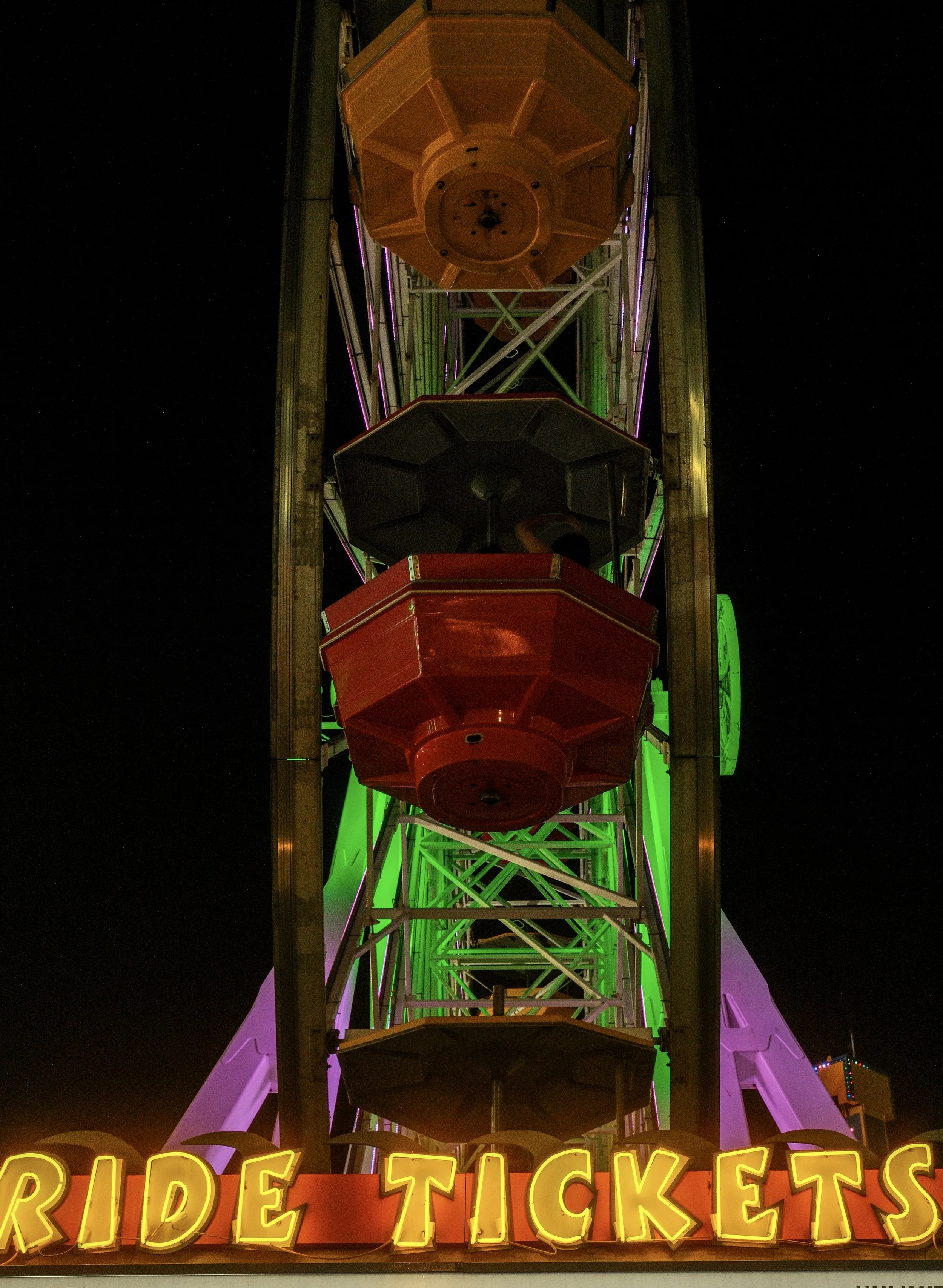 Nighttime view of a colorful Ferris wheel with illuminated green, purple, and yellow lights, and a sign reading "RIDE TICKETS" in bright yellow lights at the bottom.
