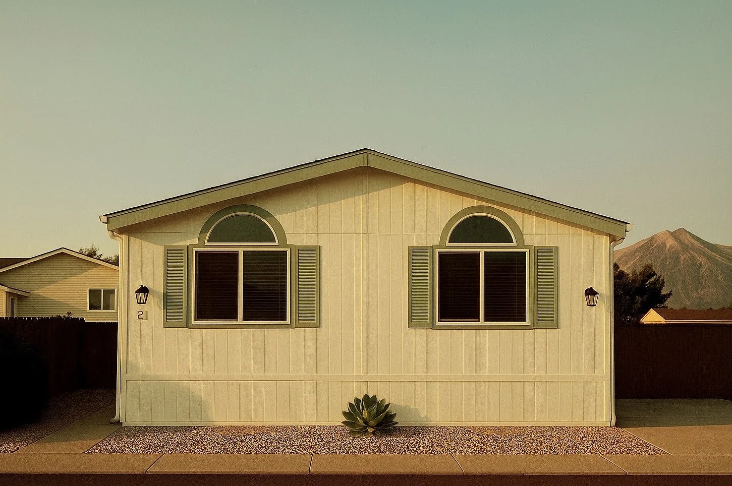 Front view of a beige house with two arched windows and green shutters, a mountain in the background, and desert landscaping in the foreground.