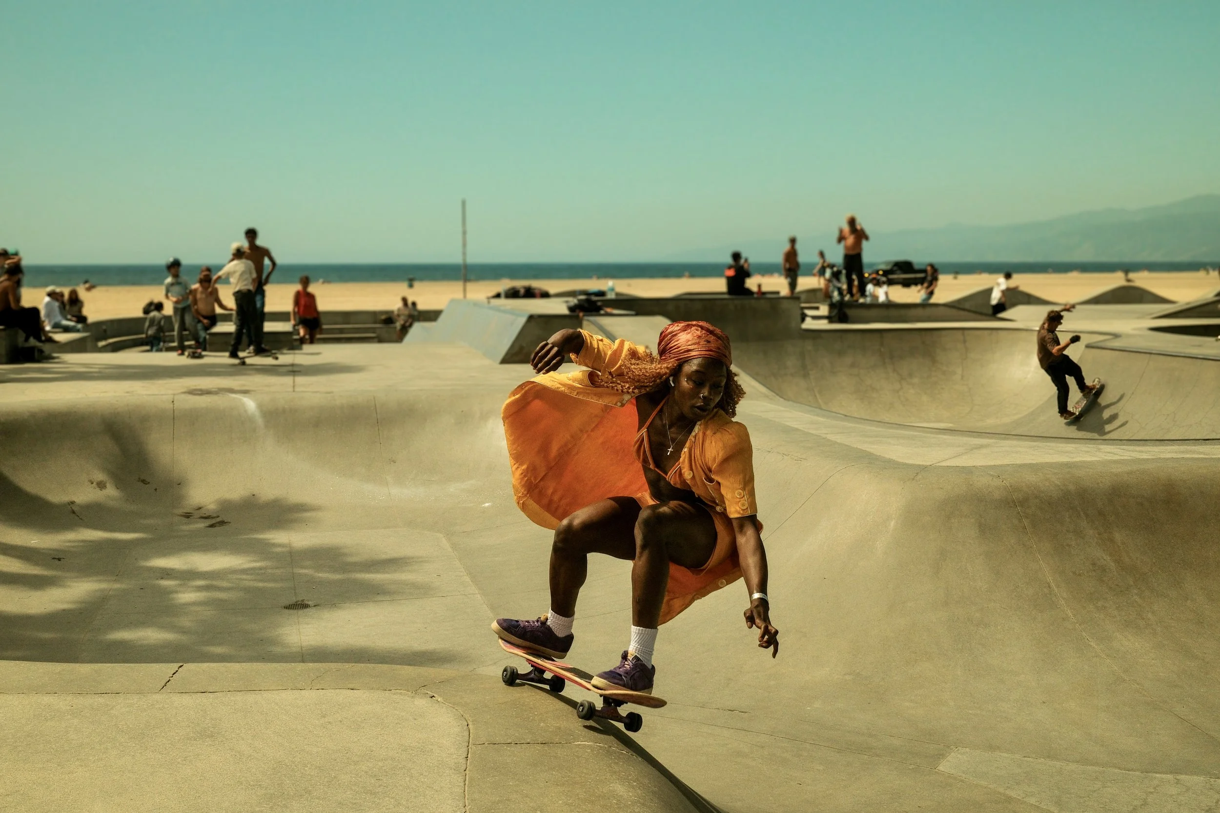 A woman skateboarding at a skatepark near the beach on a sunny day, with other people in the background skating and relaxing.