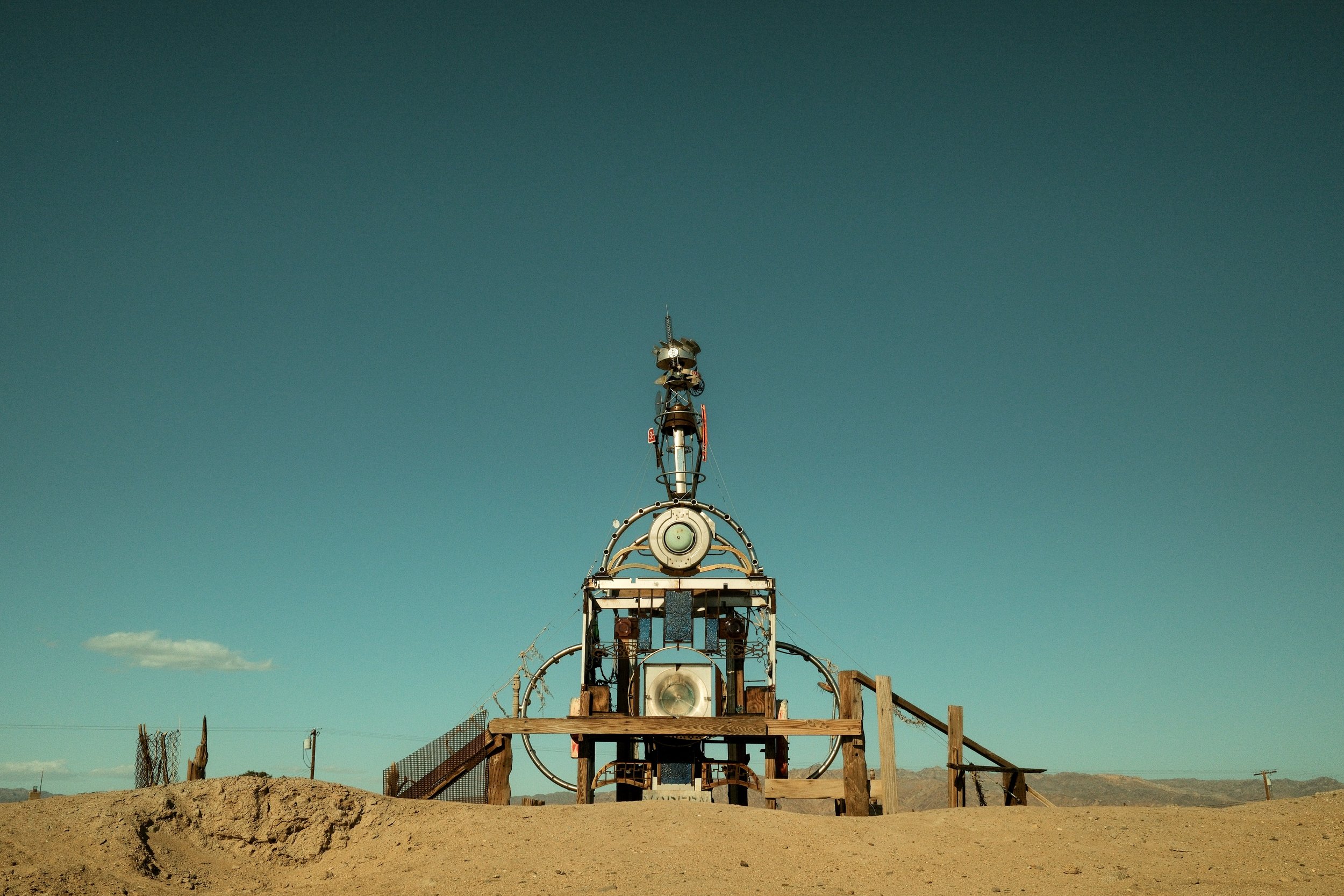 A tall metal tower with antennas and circular structures stands on a dirt mound in a desert landscape under a clear blue sky.