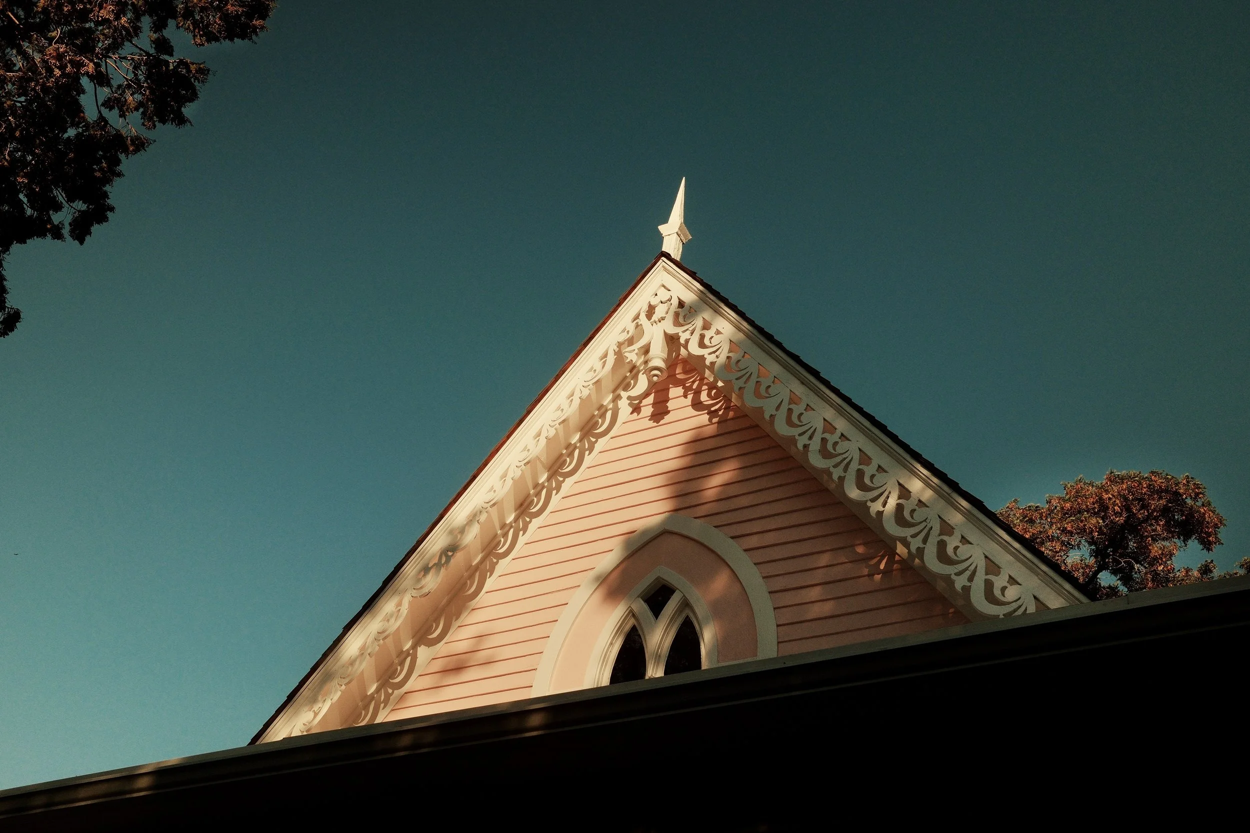 Close-up of a pink church with white decorative trim on the gable end, a pointed steeple, and a small arched window, against a clear blue sky and trees.