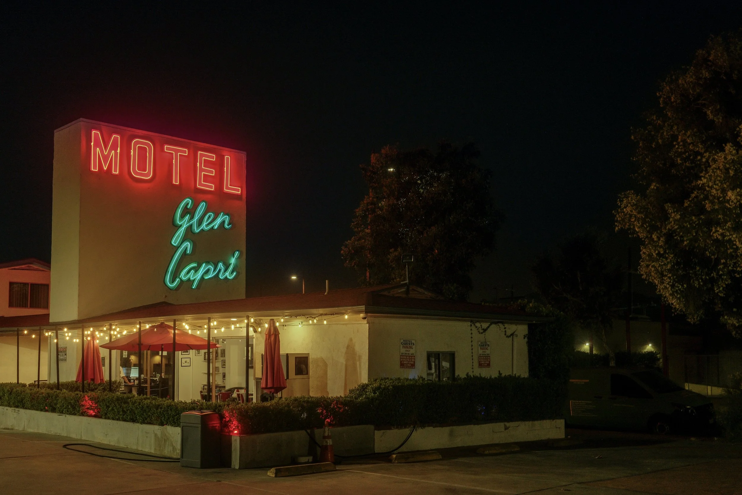 Night view of Motel Glen Capri with neon signs, outdoor seating with umbrellas, string lights, and surrounding trees and parking lot.