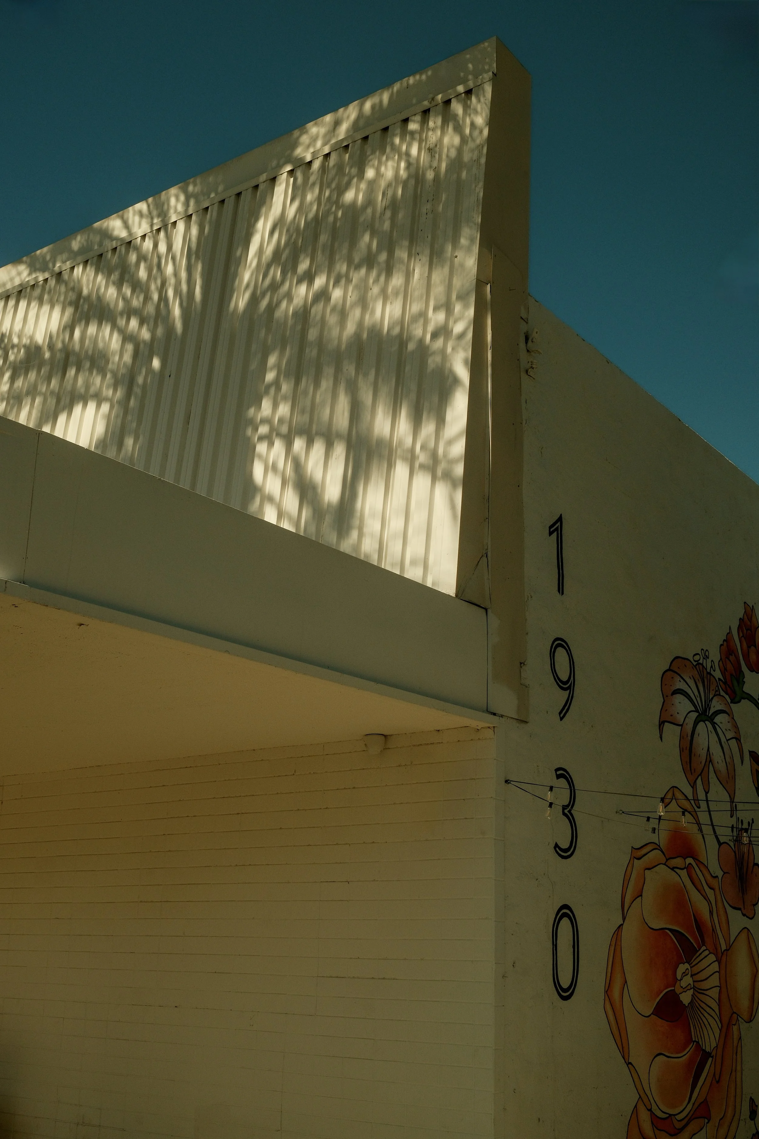 A corner of a building with painted numbers and a flower mural on the wall, featuring shadows of trees cast on the upper part, with a clear blue sky in the background.