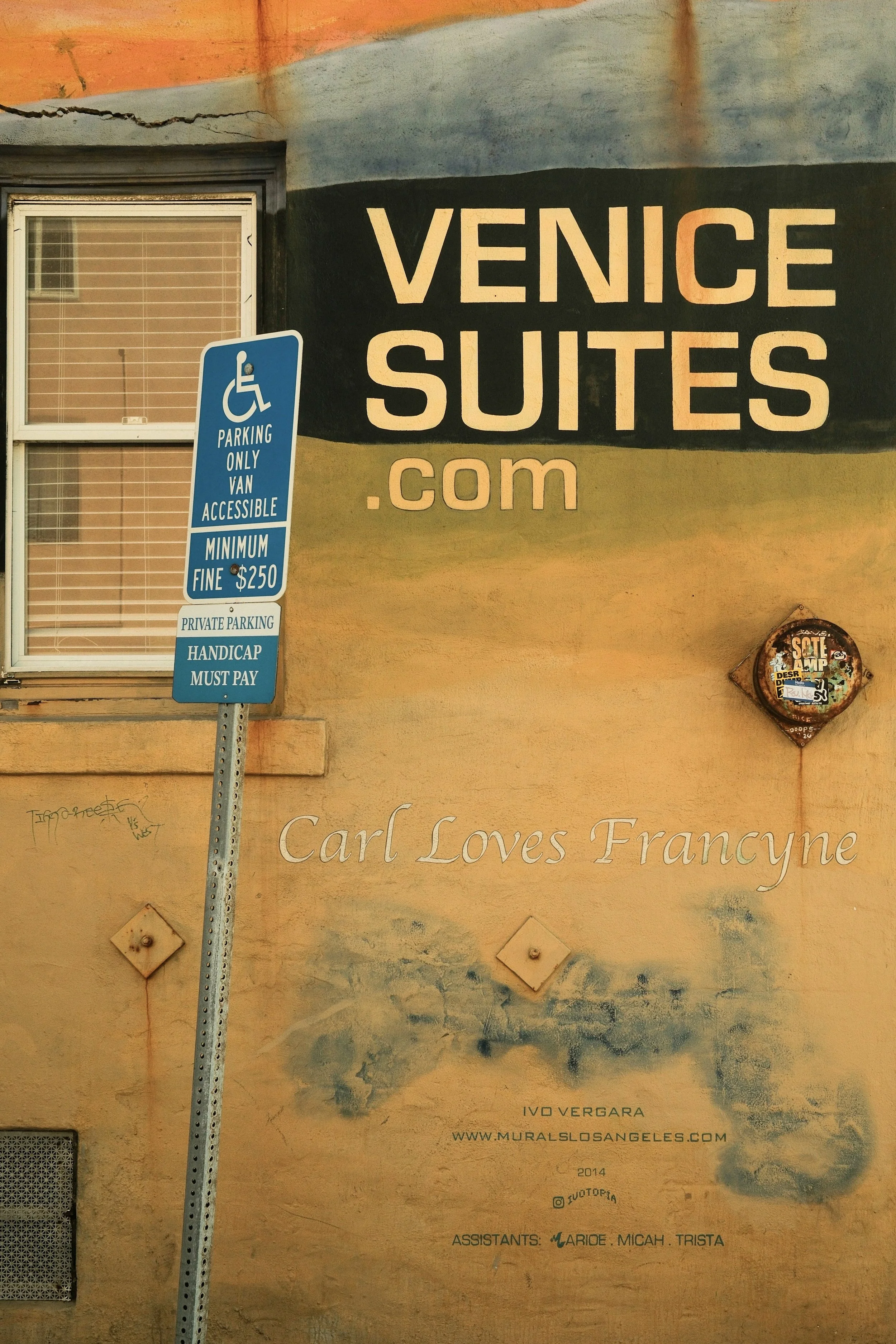 A street scene with a parking sign indicating handicapped parking with a fee of $2.50, a window with blinds, and a large mural on a wall advertising Venice Suites with the website venicesuites.com. The mural also features the phrase 'Carl Loves Francyne' along with the artist's name, Ivo Vergara, and details about the mural project.