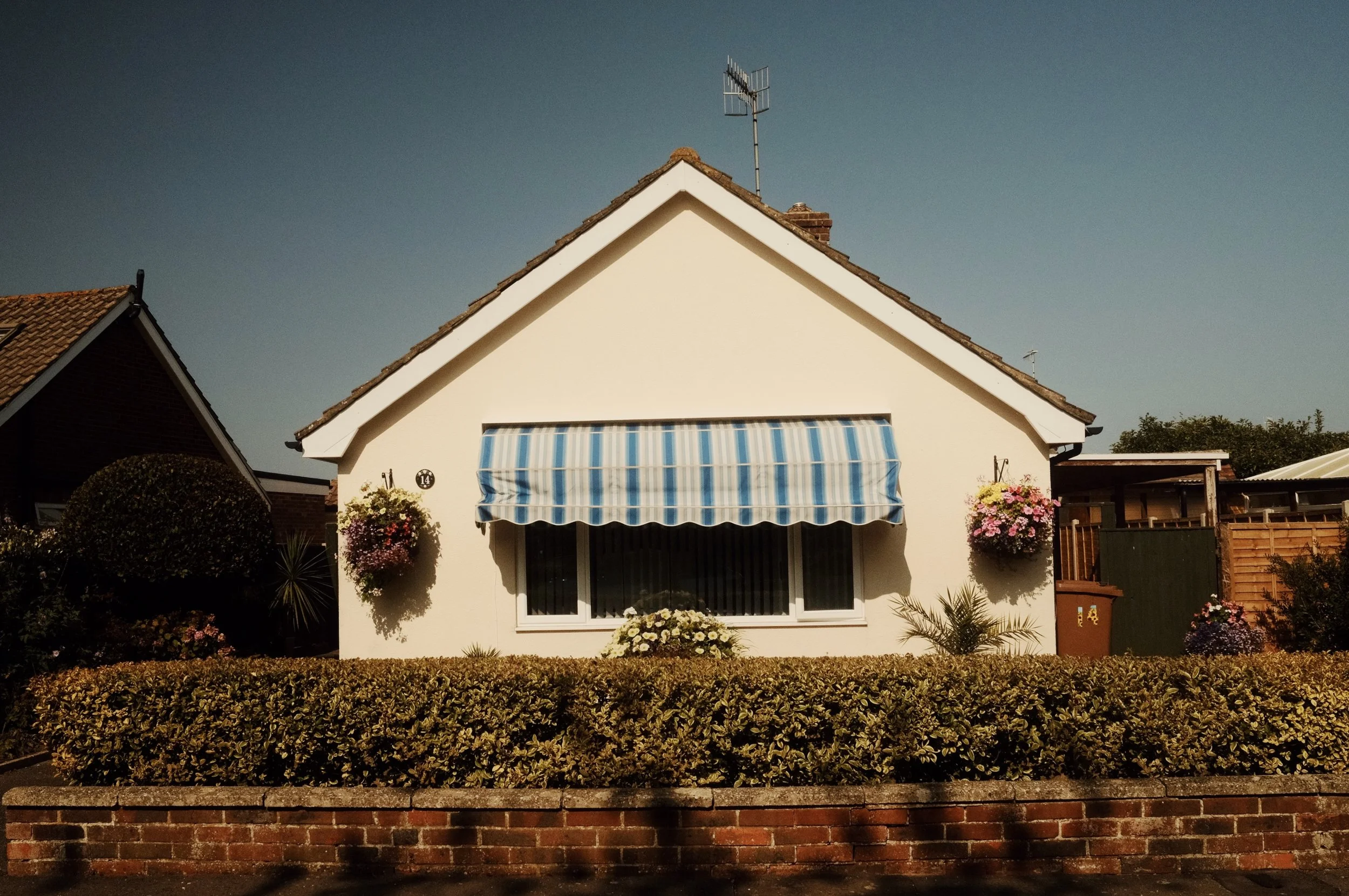 Front view of a white house with a blue and white striped awning over a window, garden with flowers, and a brick wall in front.