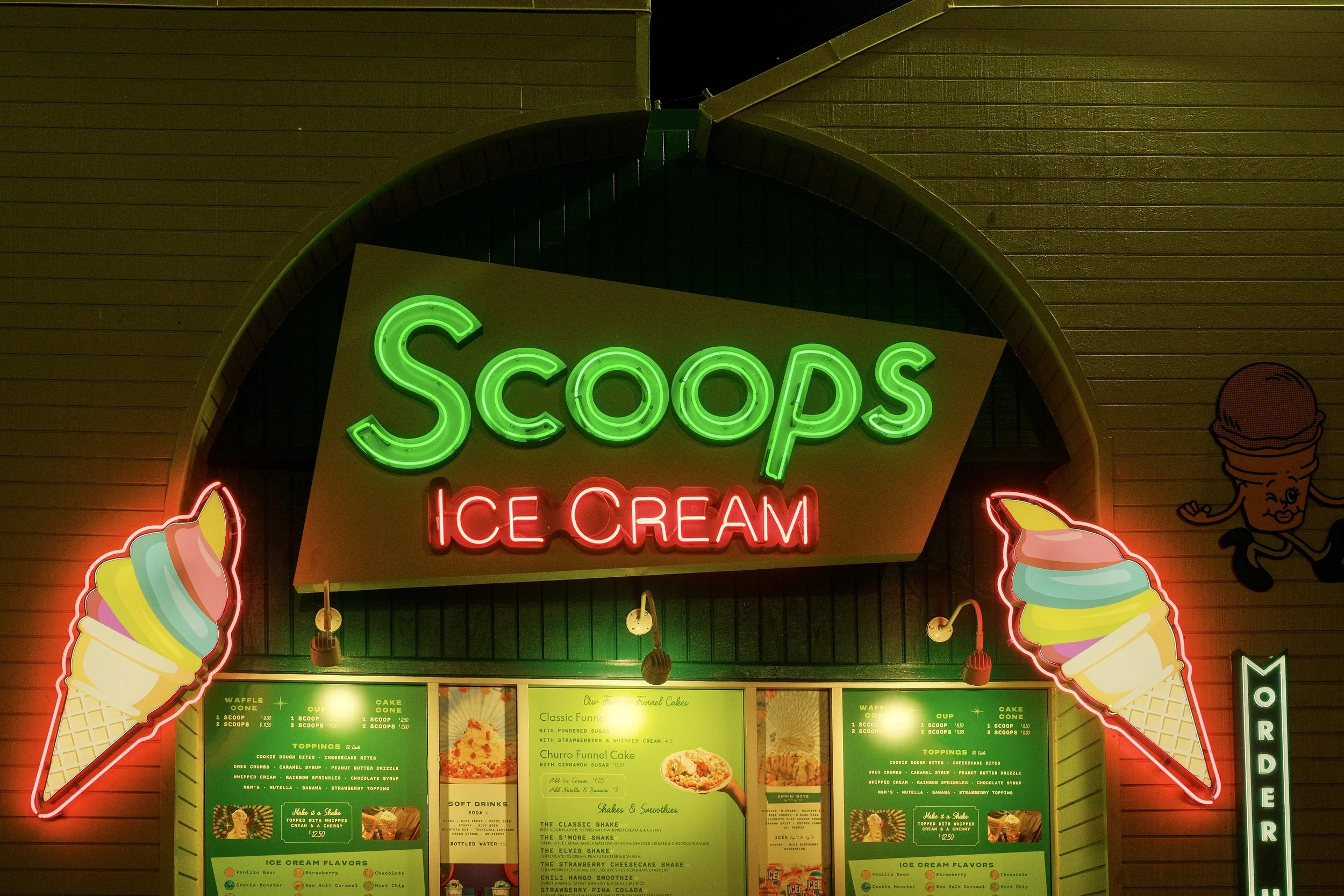 Night view of an ice cream shop with neon signs reading 'Scoops' in green and 'Ice Cream' in red, with cone illustrations on either side.