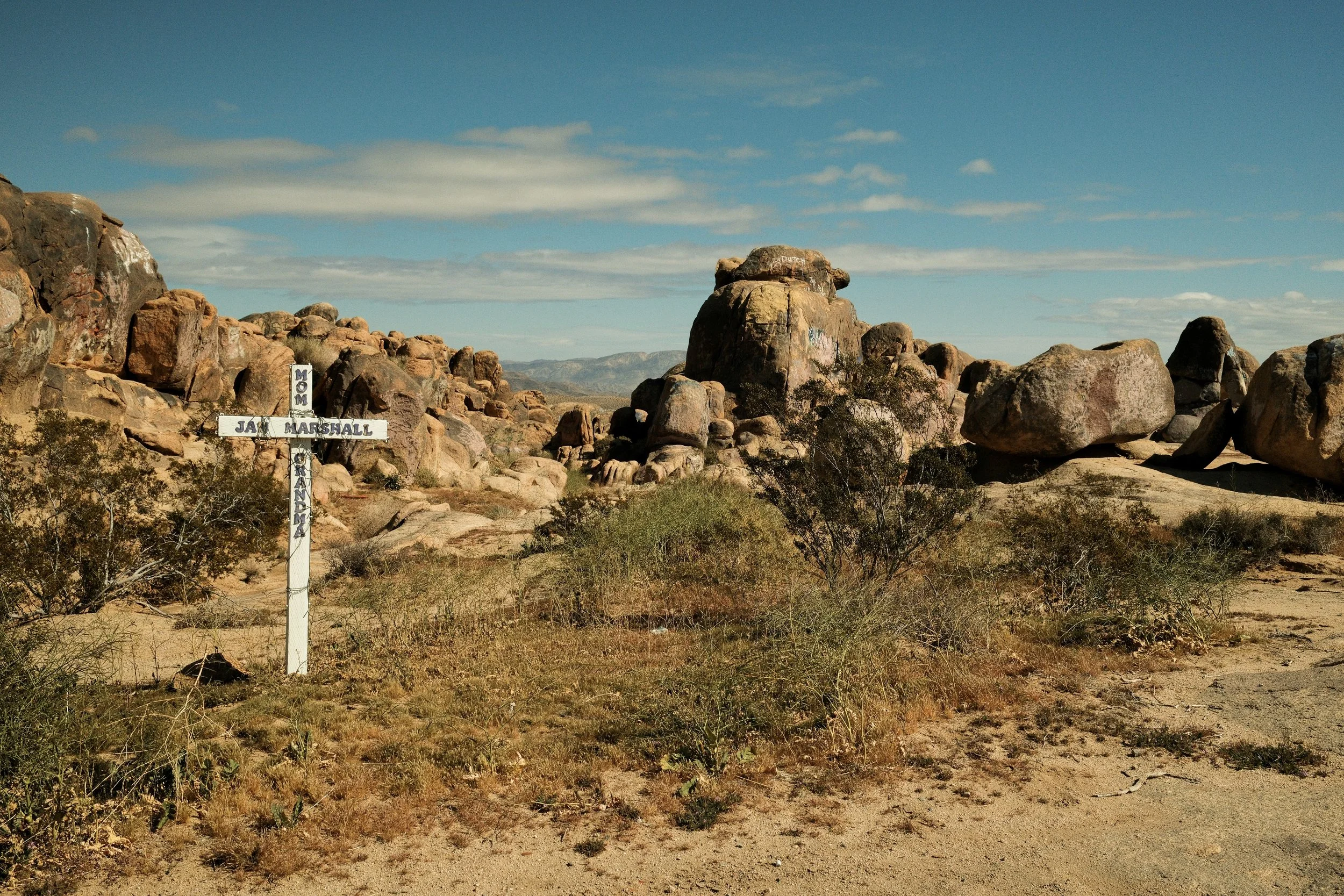 Desert landscape with large rocks and boulders, sparse vegetation, and a wooden cross with the name JAY MARSHALL, surrounded by smaller signs reading MOM, DAD, and GRANDMA. Clear blue sky with a few clouds in the background.