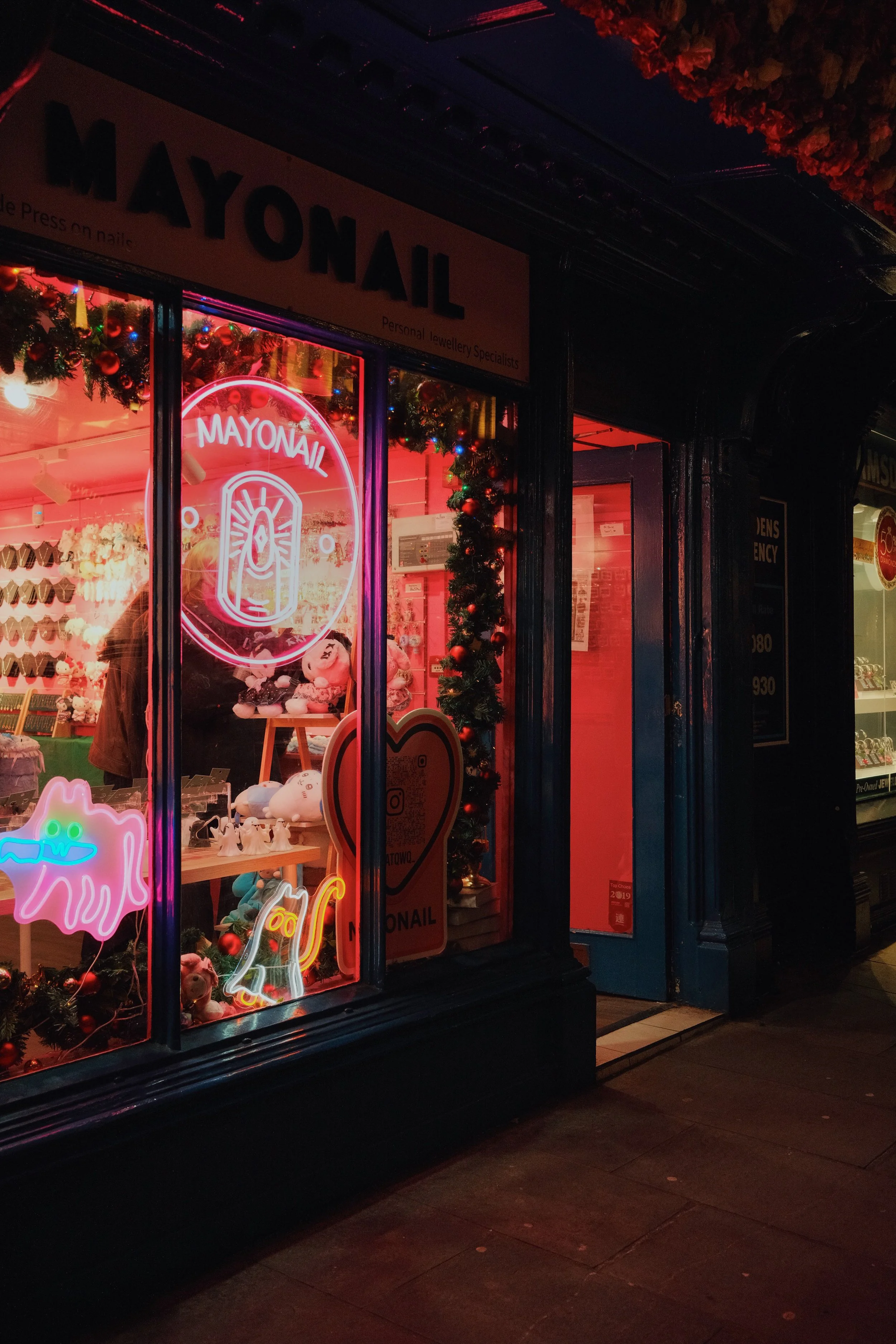 A nail salon storefront with neon signs that say 'MAYONAIL' and a stylized nail design, decorated for the holidays with Christmas garlands, ornaments, and lights, seen in a nighttime setting.