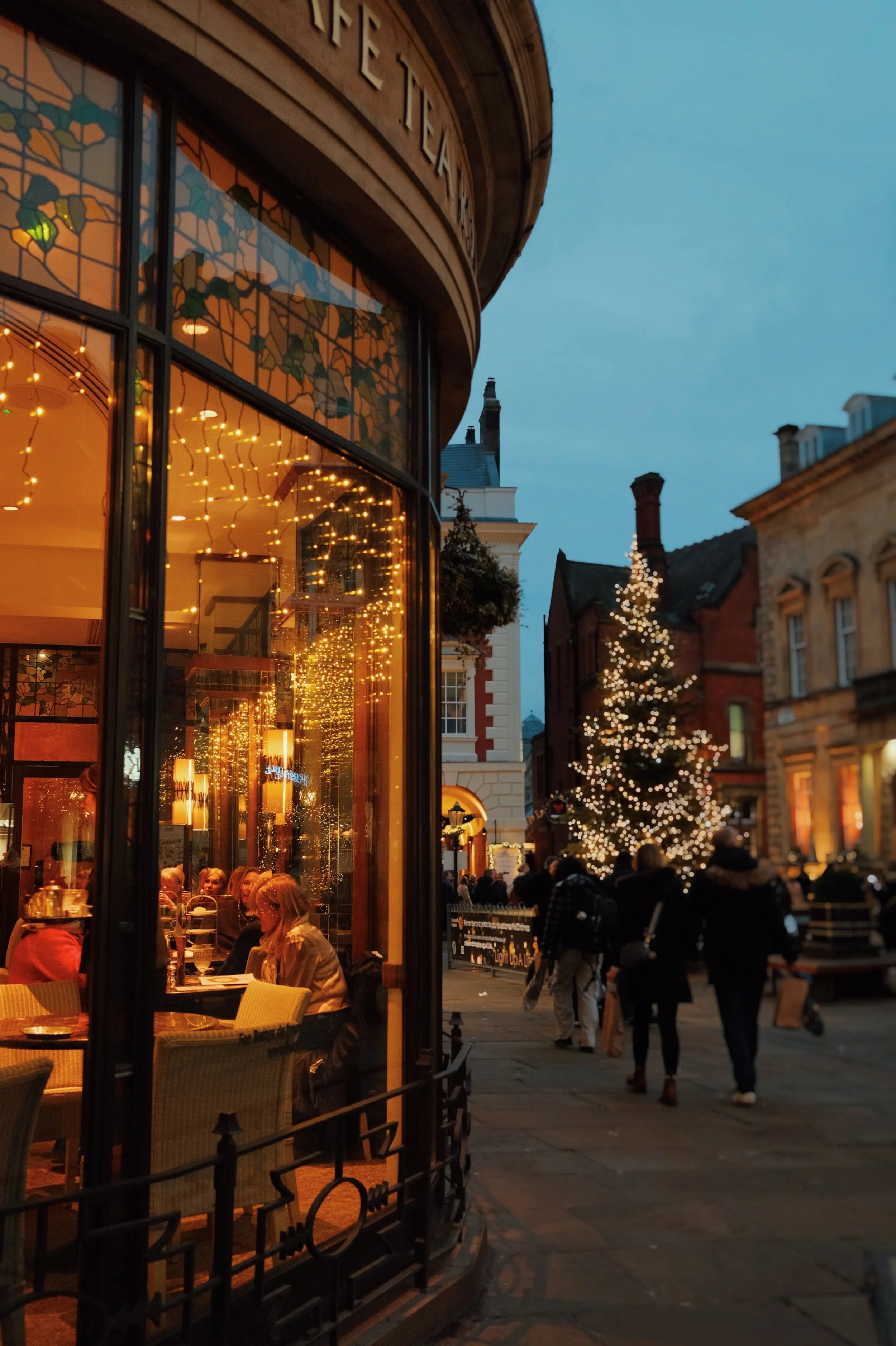 A street scene during the evening with a Christmas tree decorated with lights and people walking, viewed through the window of a restaurant with string lights inside.