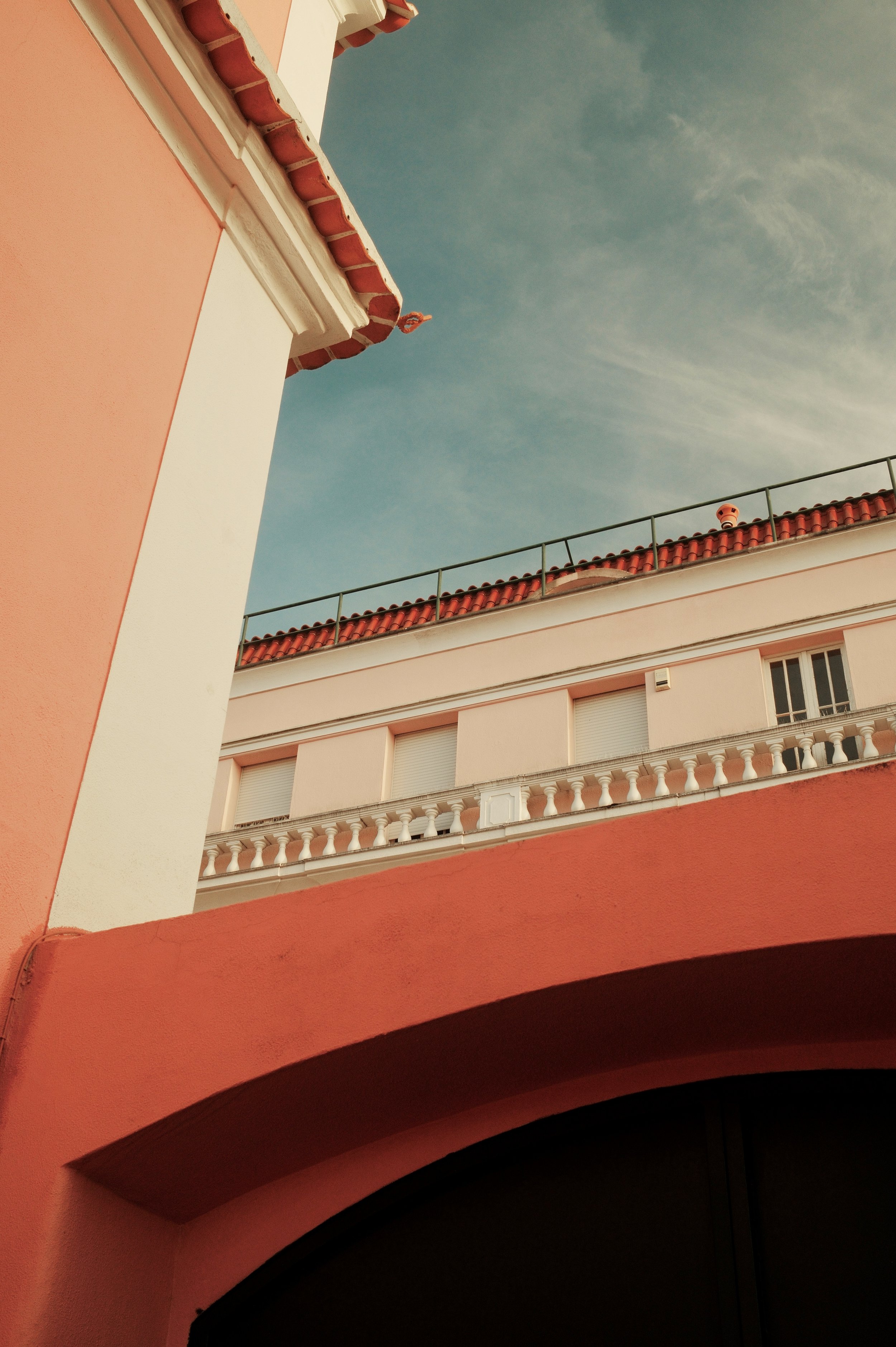 View of a pink and white building with a red-tiled roof and a balcony with balustrades, captured from below against a cloudy sky.
