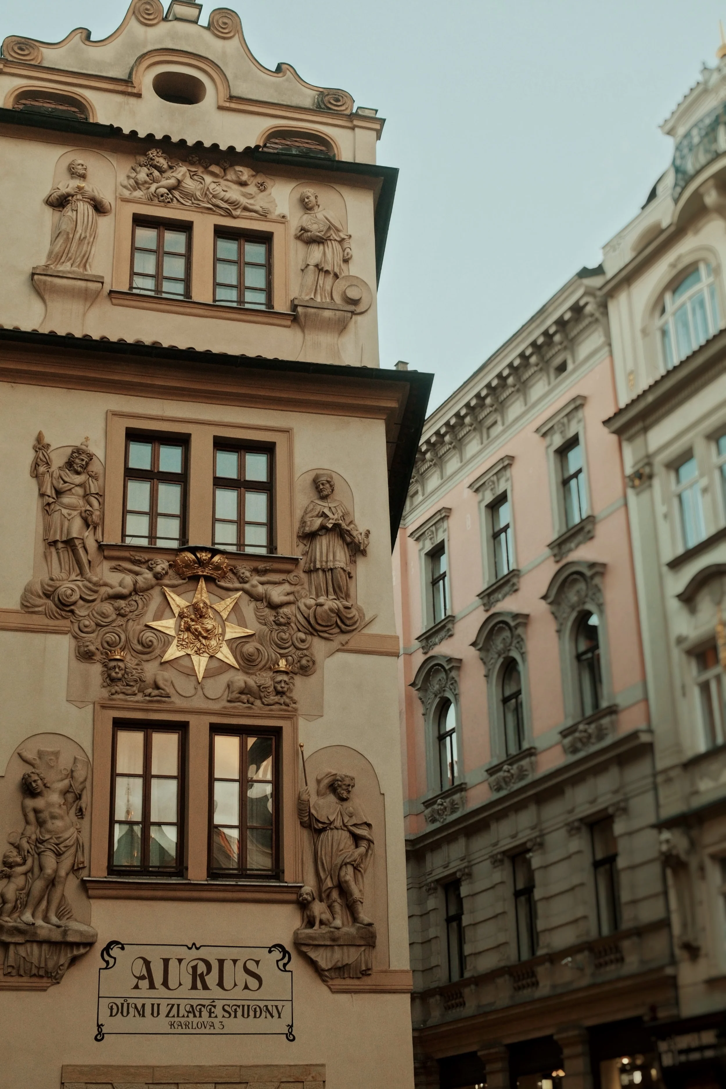 A historic European building with intricate sculptures and gold accents, with neighboring ornate buildings in a city street scene.