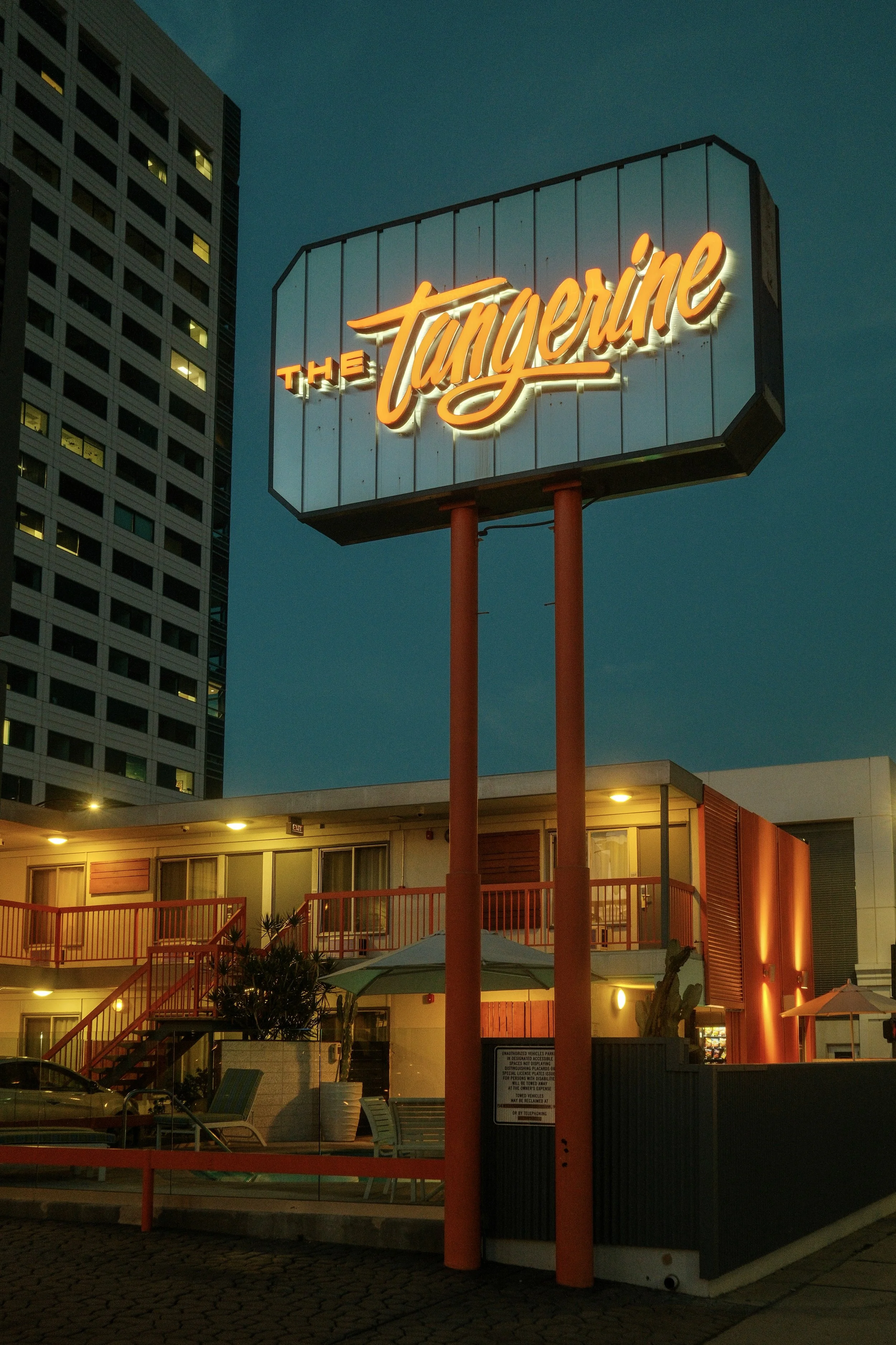 Neon sign reading 'The Tangerine' above a building with outdoor seating, umbrellas, and a parking lot, with a tall building in the background, during evening or night.
